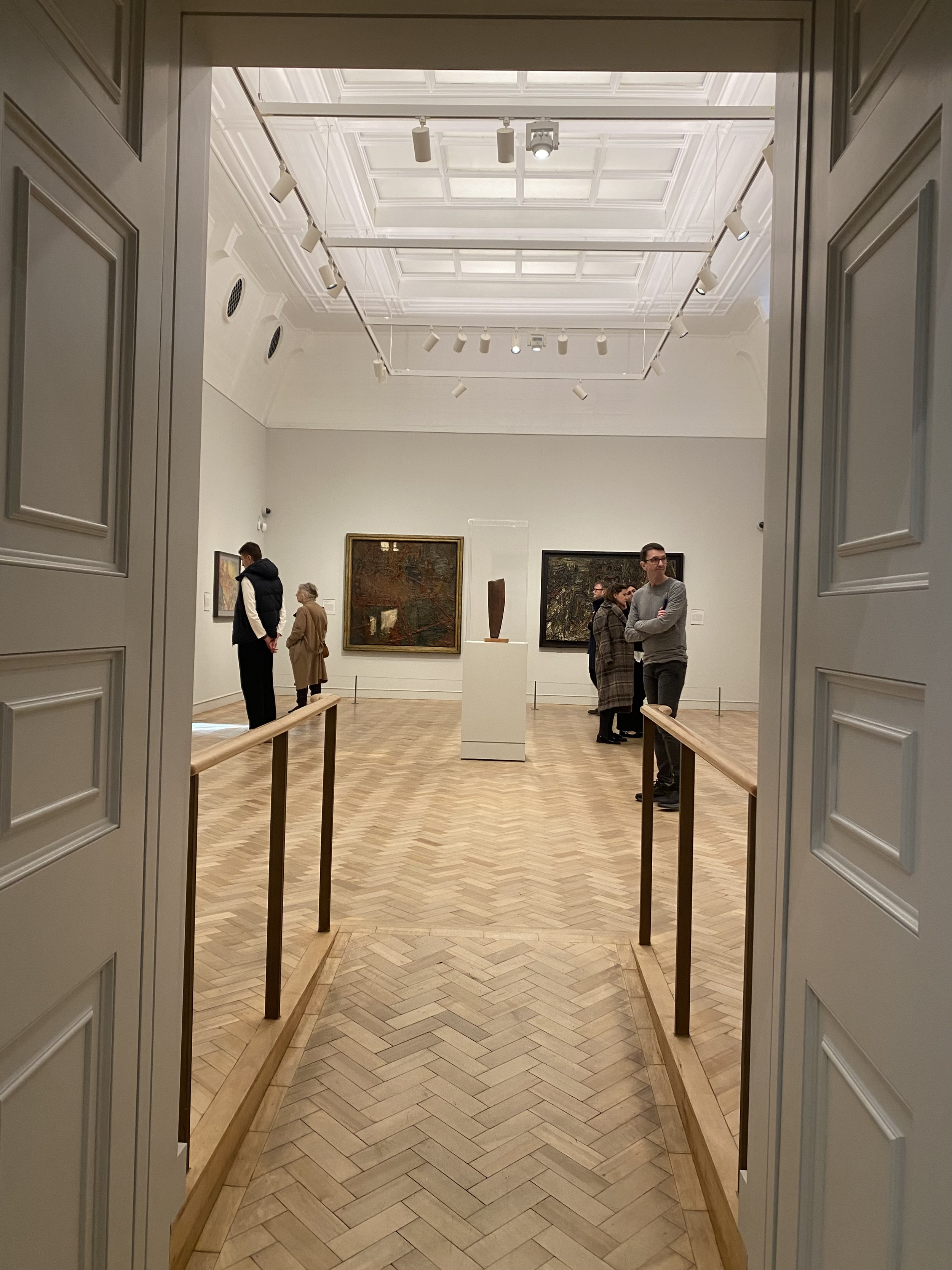 Art gallery interior with paintings on the walls and a sculpture on a pedestal. Several people are viewing the art. The gallery has a white ceiling with spotlights and wooden herringbone floor.