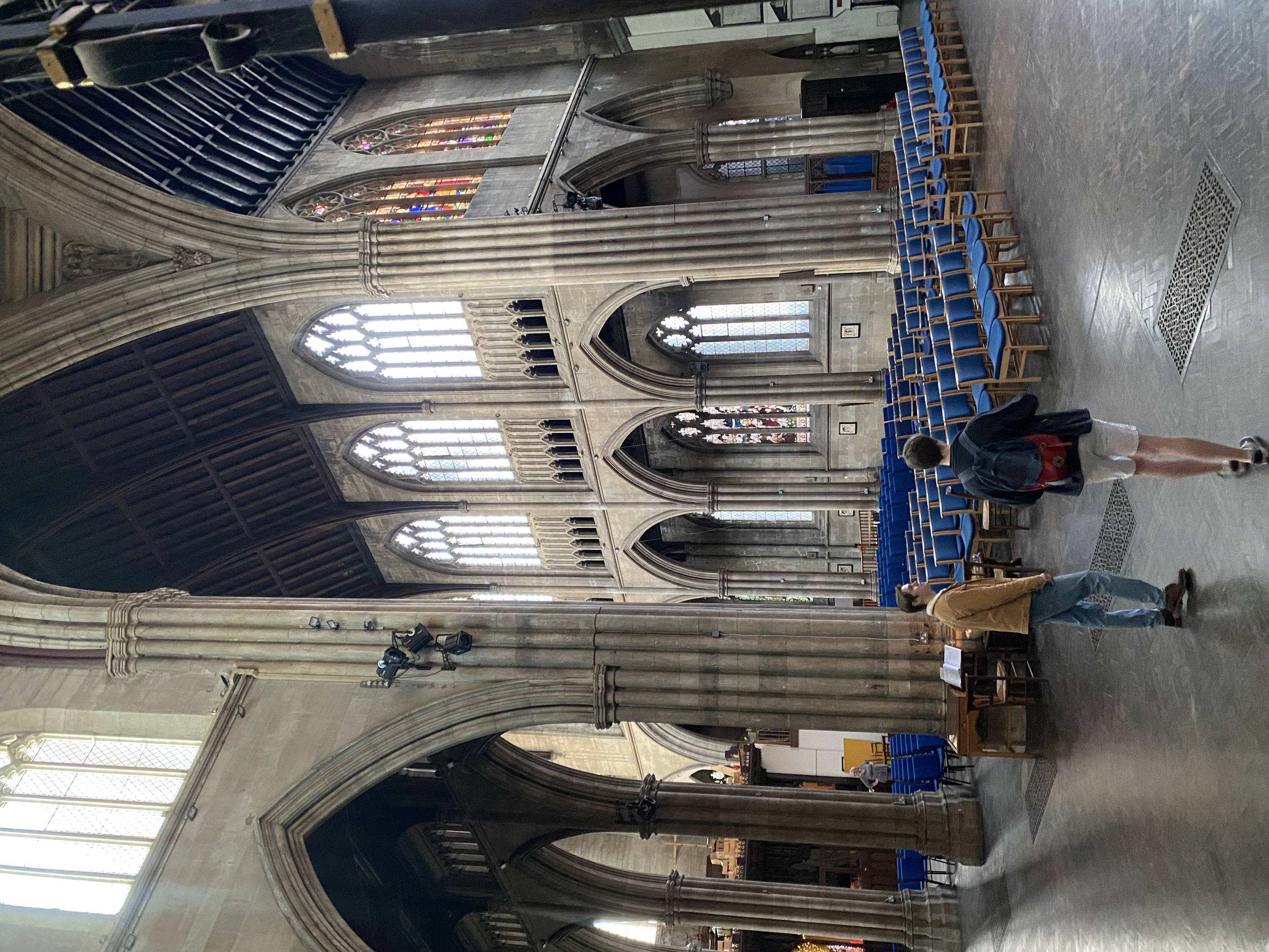 Interior of a large, historical church with Gothic architecture, featuring high vaulted ceilings, stained glass windows, and rows of blue chairs. People walking inside.