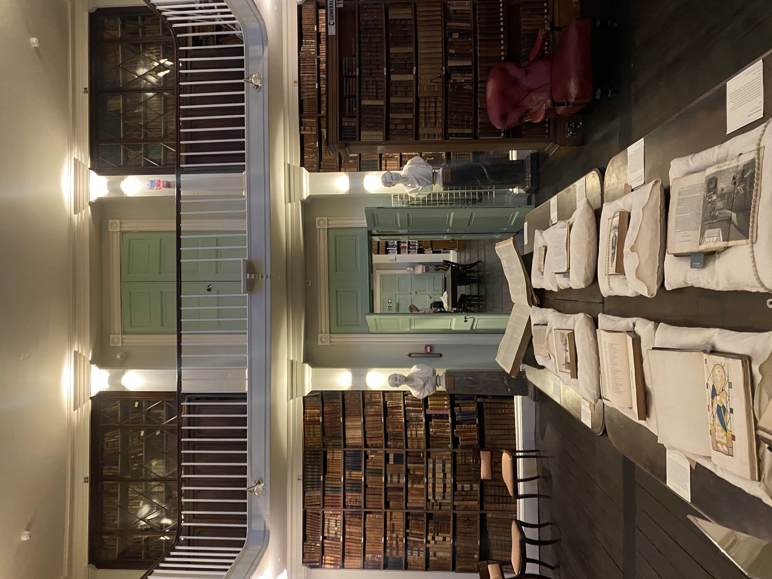 Interior of an elegant library with wooden bookshelves, open books displayed on tables, a red chair, and an upper balcony with white railings.