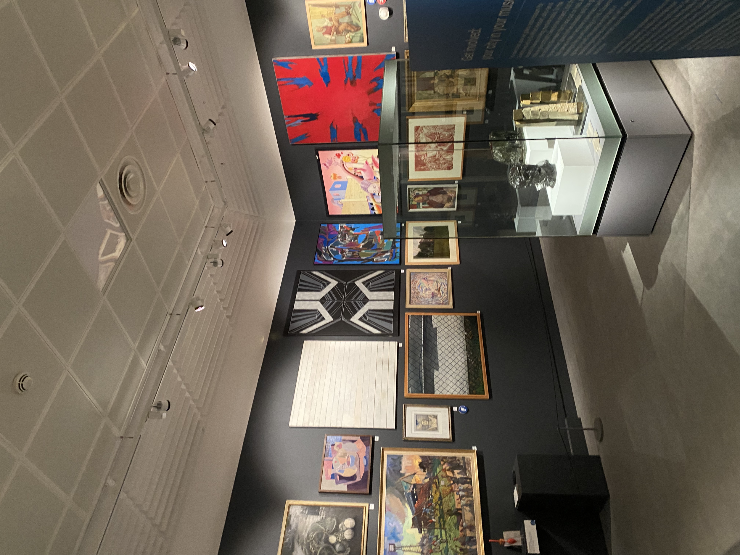 Art museum exhibition space with various framed paintings and a display case, featuring abstract and colorful artwork on black walls and spot lighting.