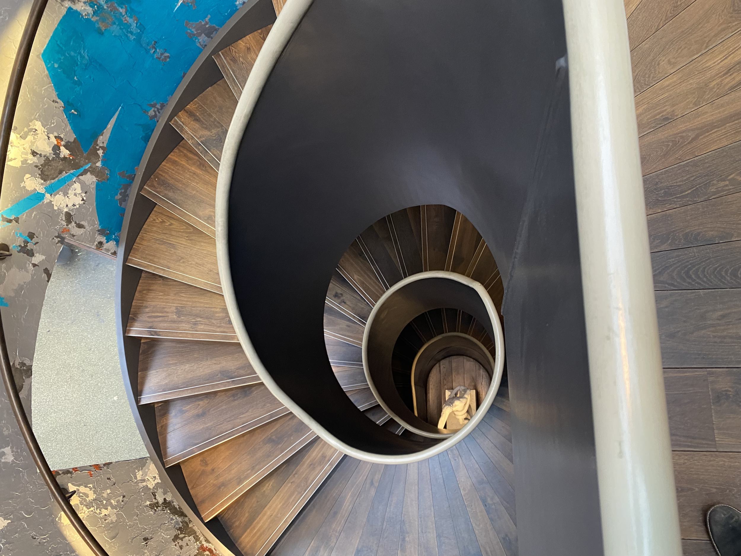 Spiral staircase with wooden steps, viewed from above, featuring a person sitting at the bottom; blue and gray wall art visible.