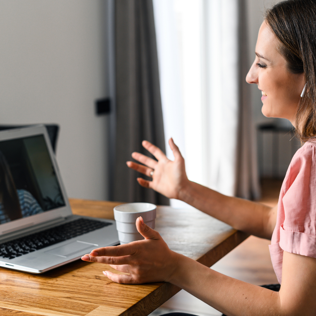 Woman talking on a video call from her home office, gesturing with hands, sitting at a wooden table with a laptop and a white mug.