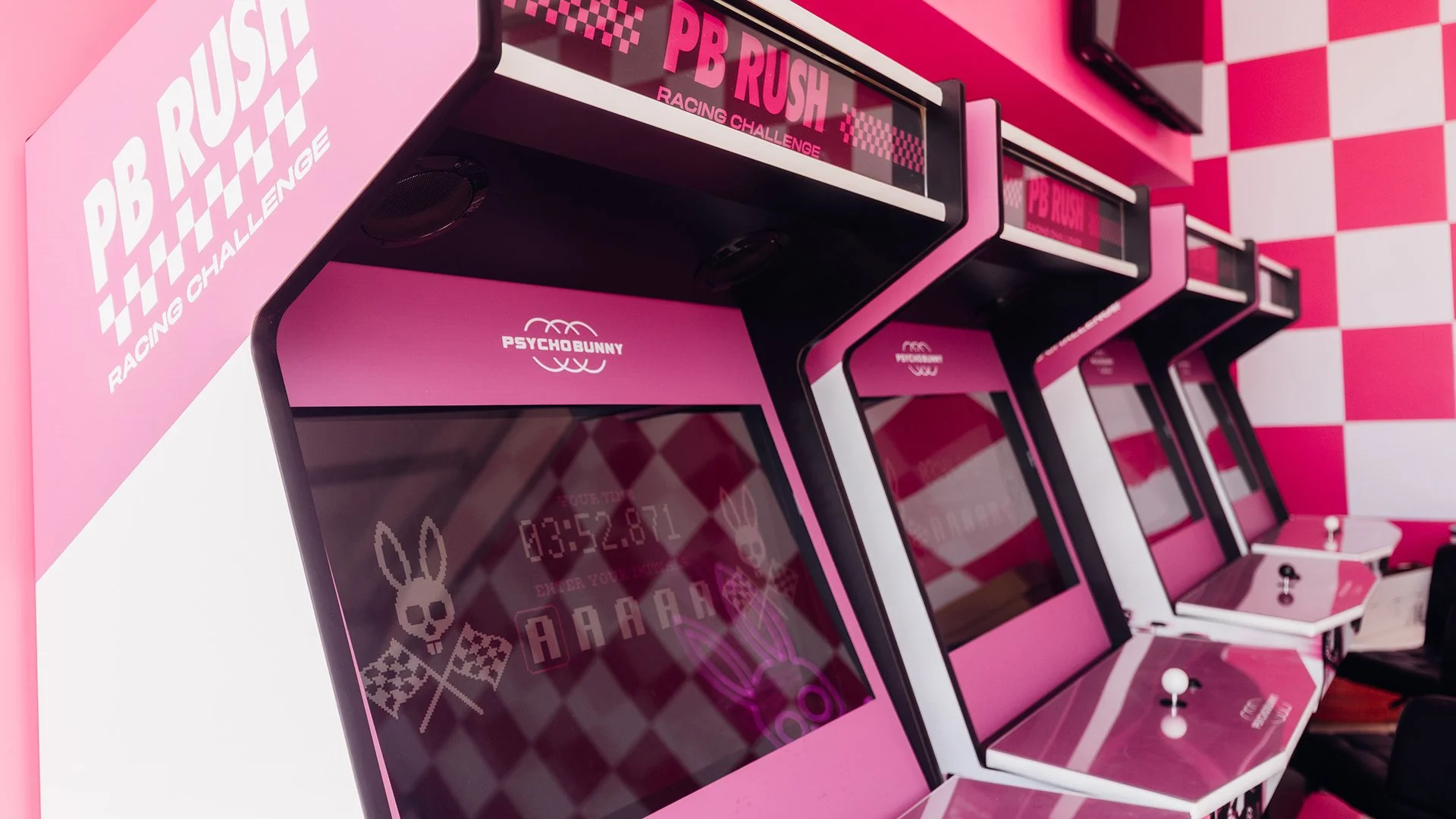 Four identical PB Rush arcade cabinets featuring Jude Buffum’s custom Psycho Bunny racing video game at the Psycho Bunny F1 Miami Grand Prix event, surrounded by pink lighting and checkered racing graphics.