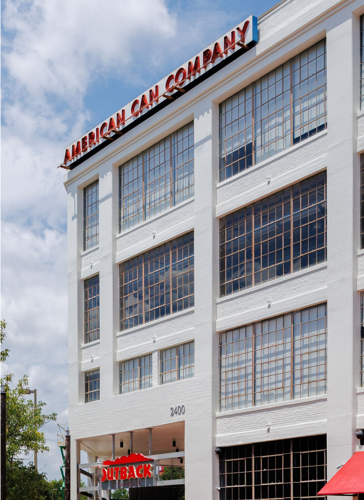 White multi-story building with large windows, displaying signs for American Can Company and Outback Steakhouse, with a partial view of a blue sky with clouds.