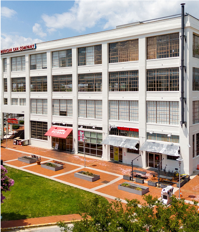 Four-story white building with large windows, signs indicating a pancake house, pizza, and Outback Steakhouse, outdoor seating area with tables and chairs, brick walkway, landscaped grass area with planters, and blue sky with some clouds.