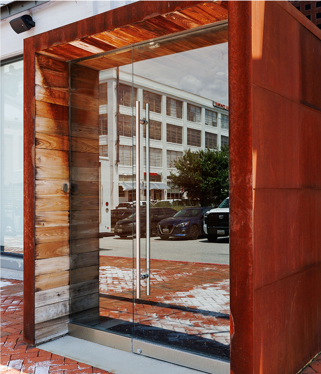 A modern storefront entrance with glass double doors, surrounded by wood and rust-colored metal panels, reflecting a city street with parked cars and a white building across the street.