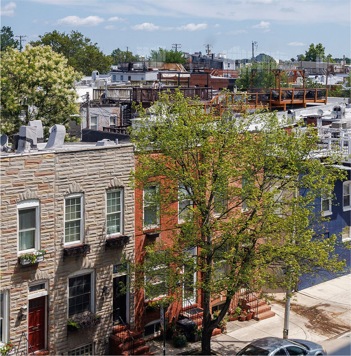 Cityscape with residential buildings, a large tree in the foreground, and a clear blue sky with some clouds.