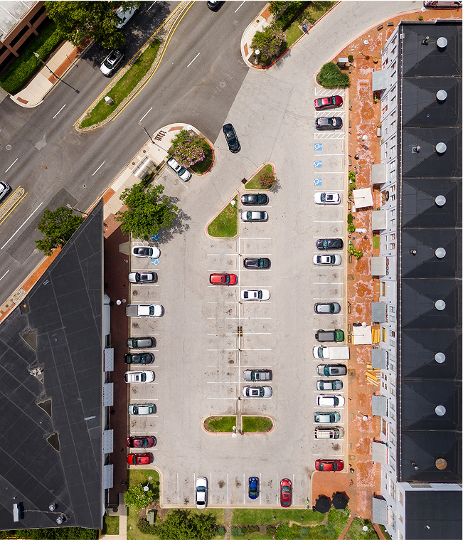 An aerial view of a parking lot with several parked cars and surrounding buildings, trees, and sidewalks.