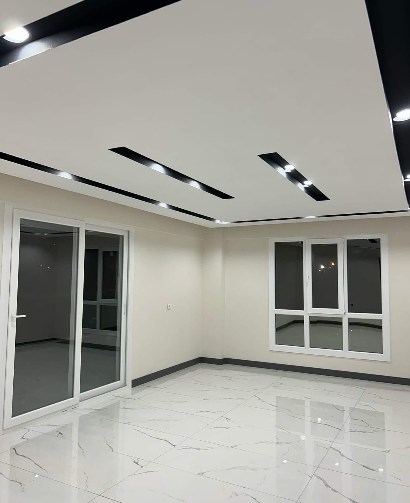 Ein leerer moderner Raum mit glänzendem weißem Fliesenboden, großen Fenstern und abgehängter Decke mit eingebauten Lichtern.