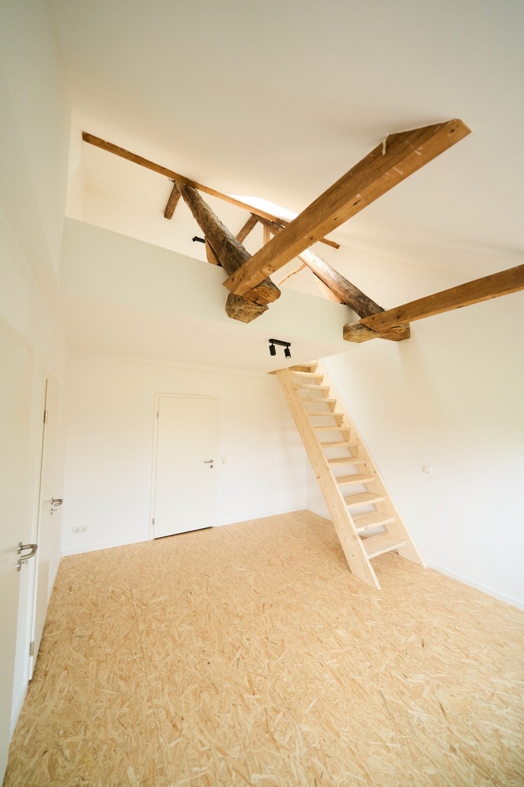 Minimalistisch gestalteter Raum mit Holztreppe zu einem offenen Dachboden und sichtbaren Holzbalken.