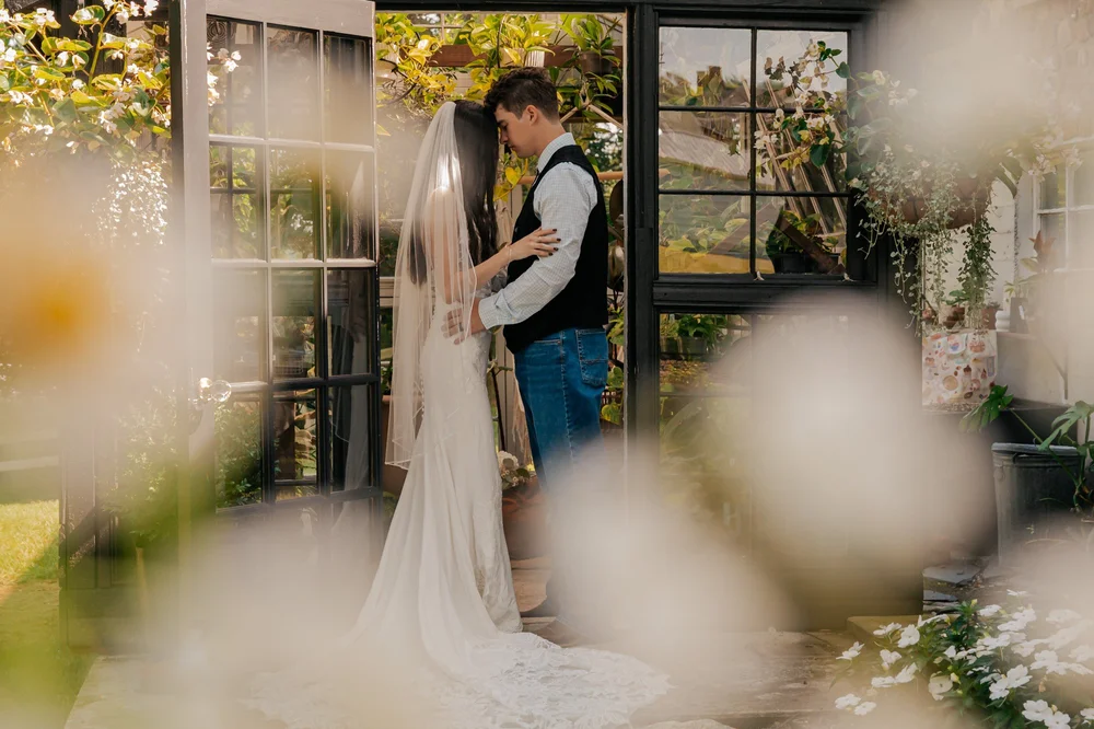 A bride and groom standing close and touching foreheads inside a greenhouse surrounded by plants and flowers