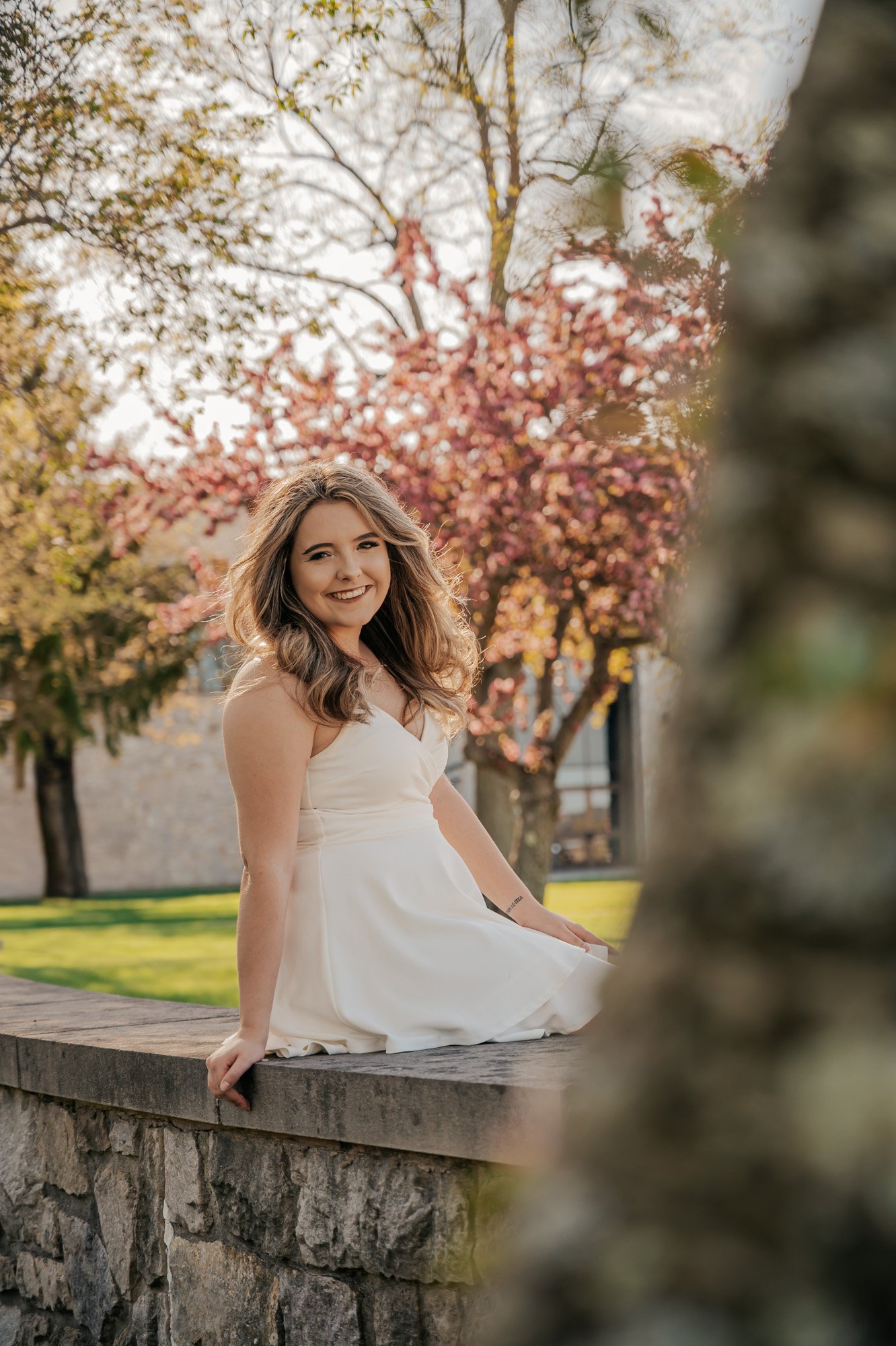 Woman in a white dress sitting on a stone wall with blooming trees in the background.