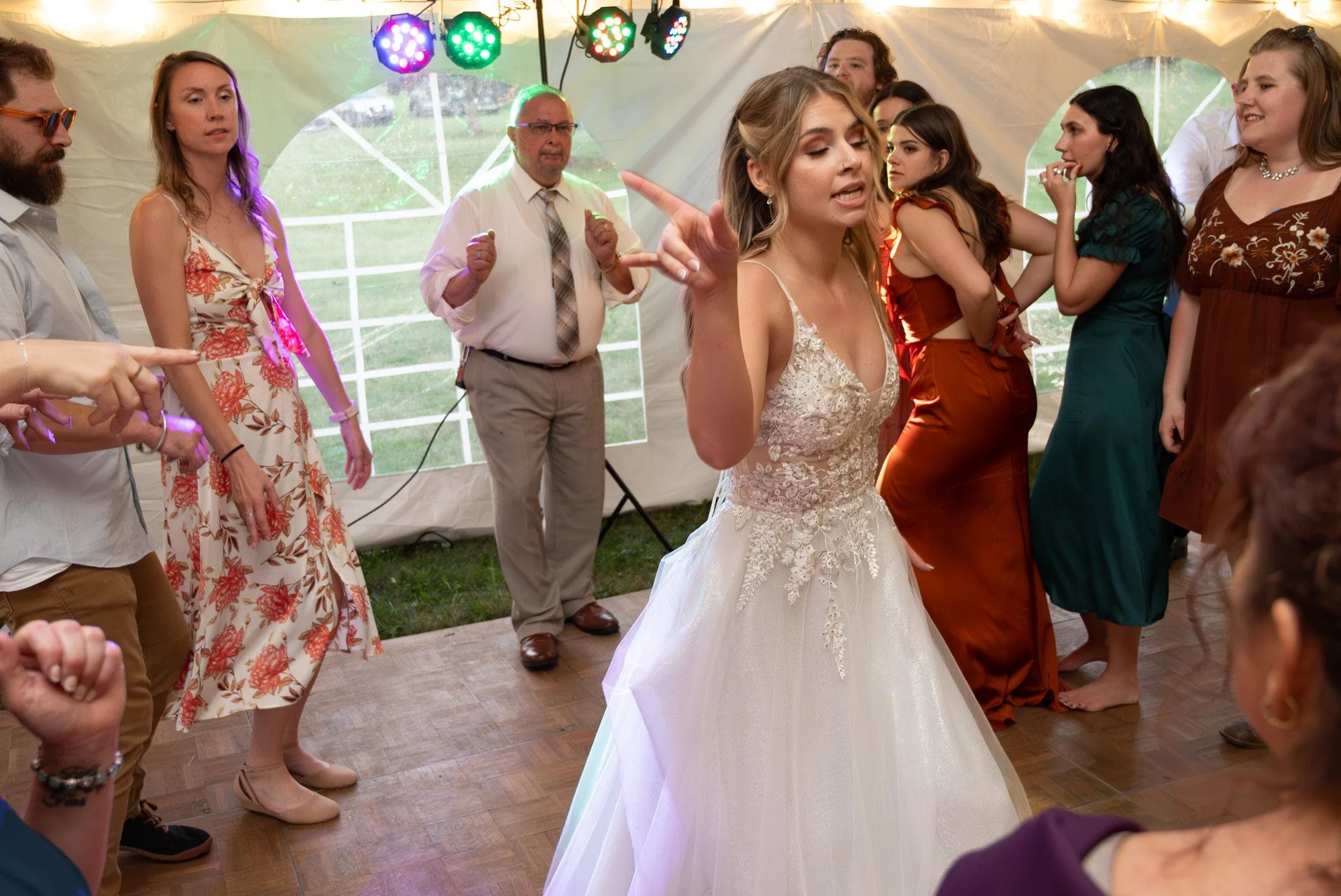 A bride in a white wedding dress is dancing with her friends and family at a wedding reception inside a tent, with colorful lights overhead.