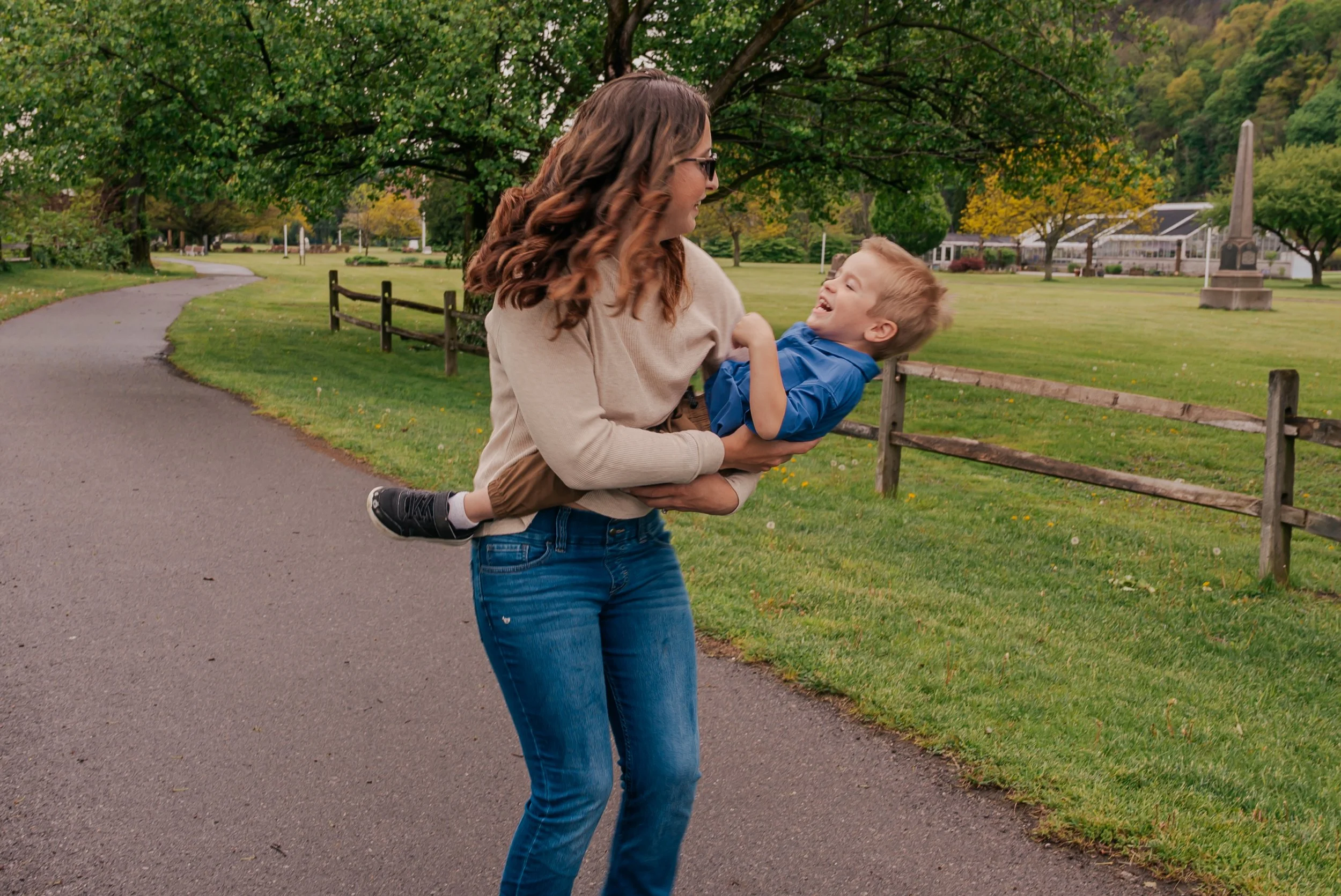 A woman with wavy brown hair, wearing sunglasses, a beige sweater, and blue jeans, holding a laughing young boy in a blue shirt and brown pants in her arms in a park with green grass, trees, a path, and a monument in the background.