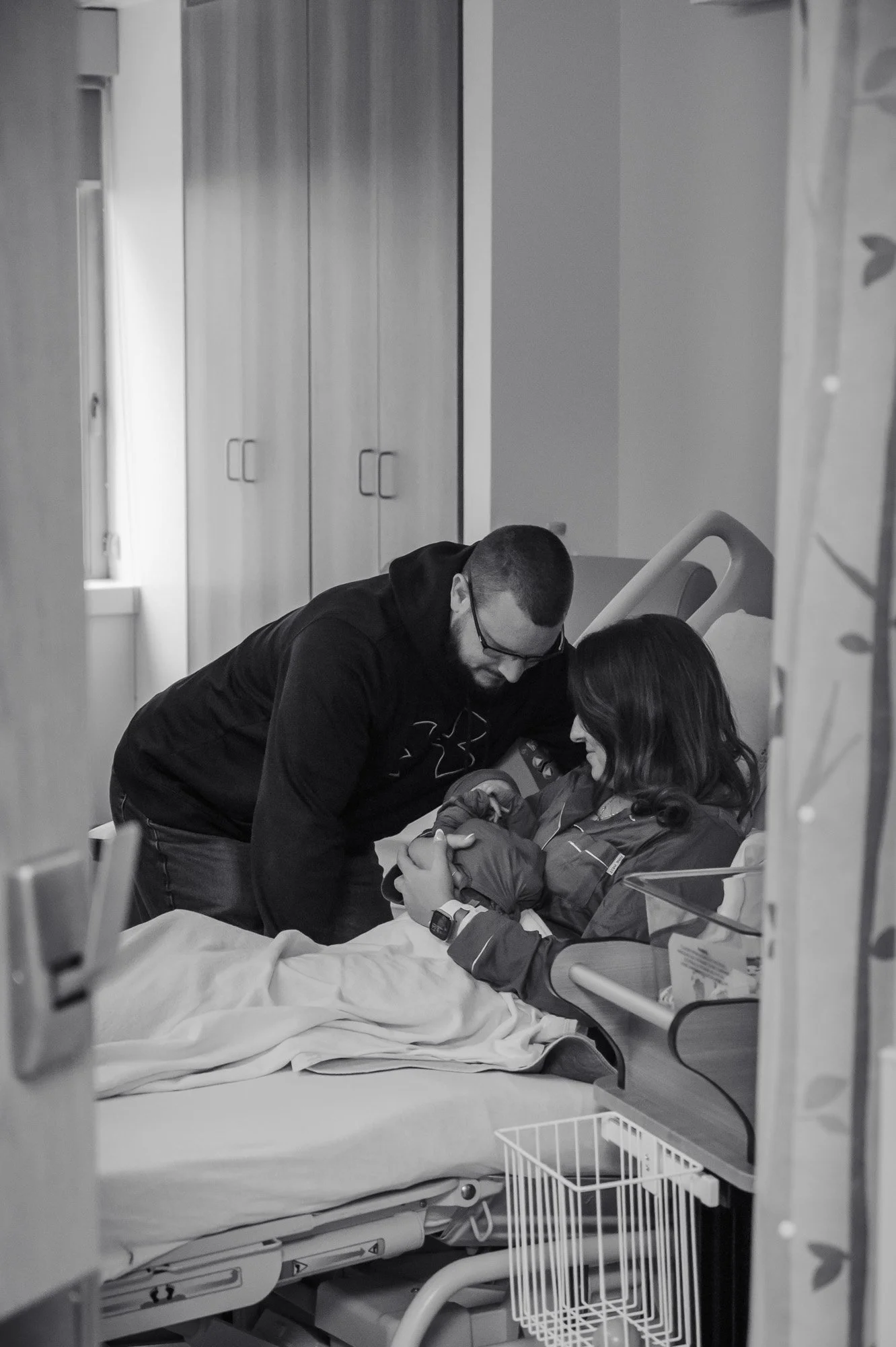 A man with glasses leans over a woman sitting in a hospital bed, holding her hand and possibly talking to her, in a hospital room.