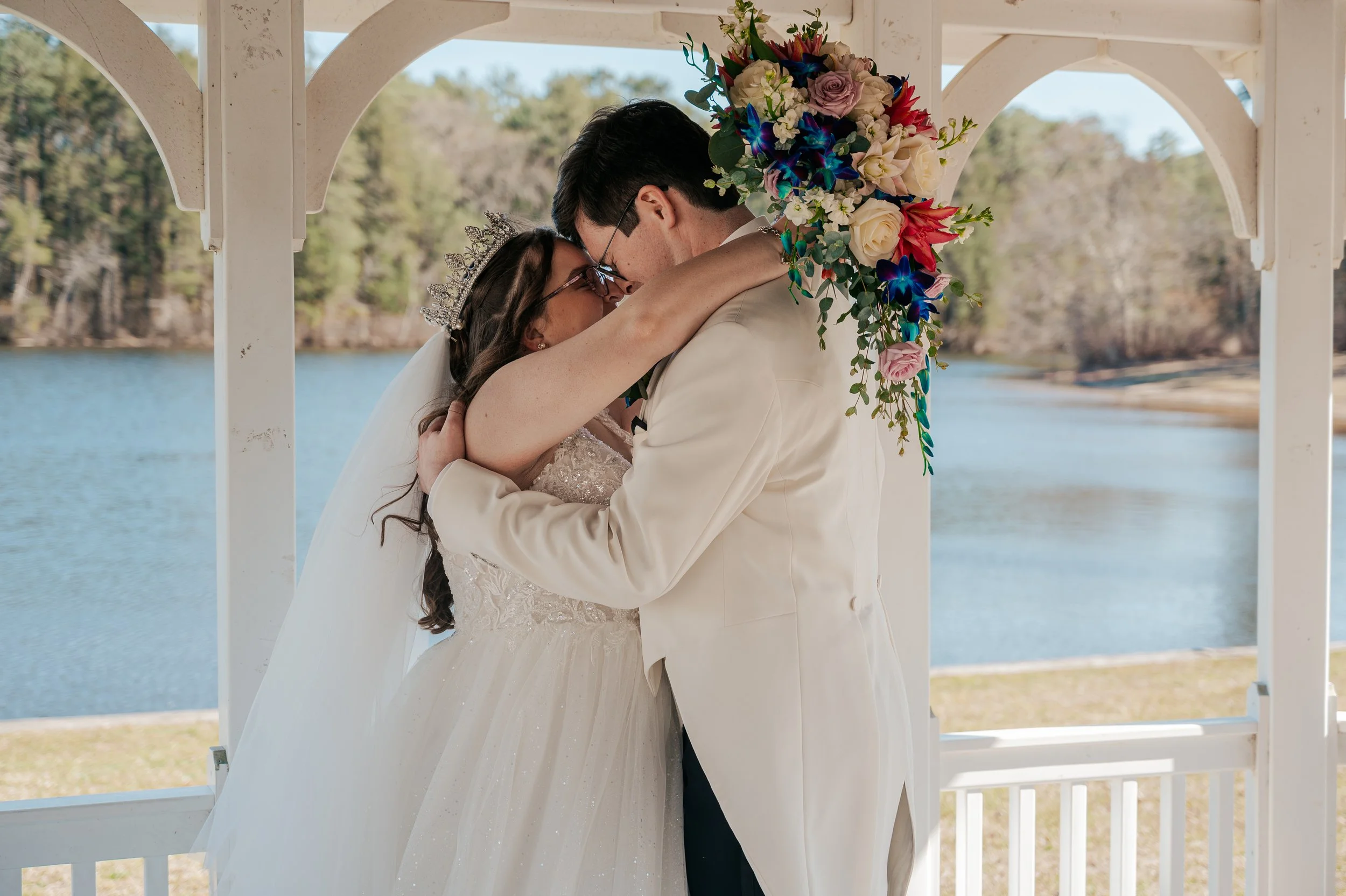 A bride and groom sharing a kiss outdoors under a white gazebo with a river and trees in the background, the bride wearing a crown and a white wedding gown, and the groom in a beige suit with a large floral bouquet on his shoulder.  Pennsylvania elop