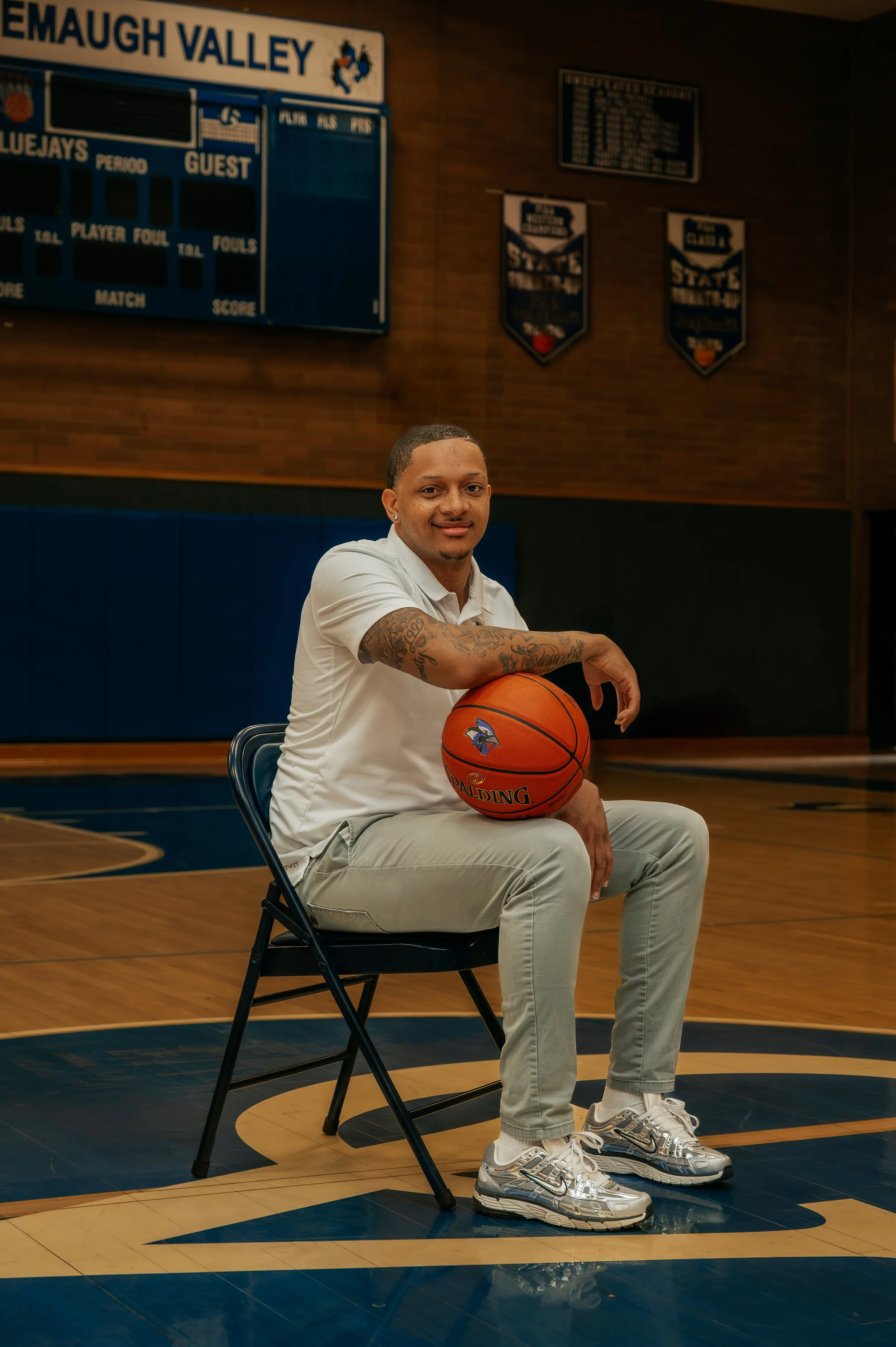 Man sitting on a chair in a basketball gymnasium holding a basketball, wearing a white polo shirt, light pants, and metallic sneakers.