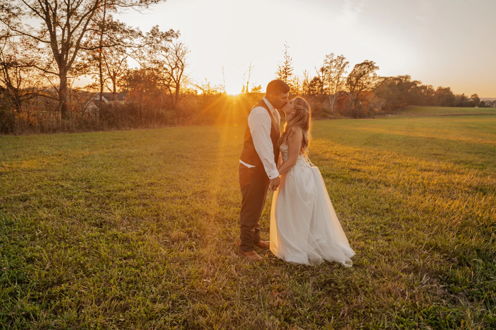 A newlywed couple kissing in a grassy field at sunset, holding hands, with trees and a distant horizon in the background.