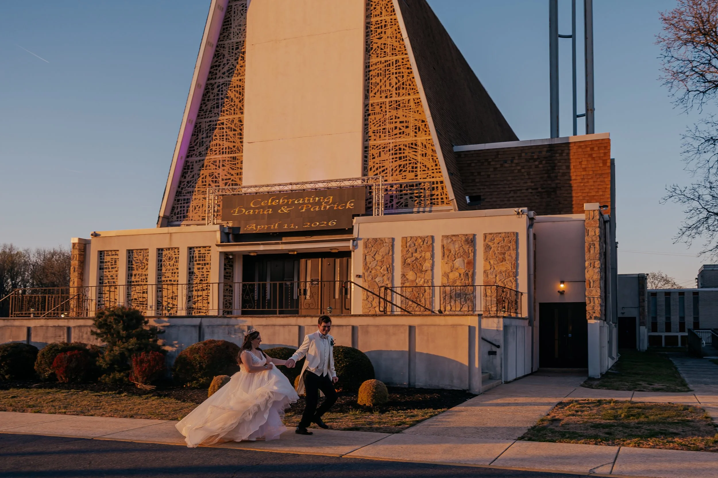 A bride and groom walking hand in hand outside a church at sunset during their wedding. The church has modern architecture with intricate lattice detailing and a banner displaying 'Celebrating Dana & Patrick April 11, 2026'. The bride is wearing a wh