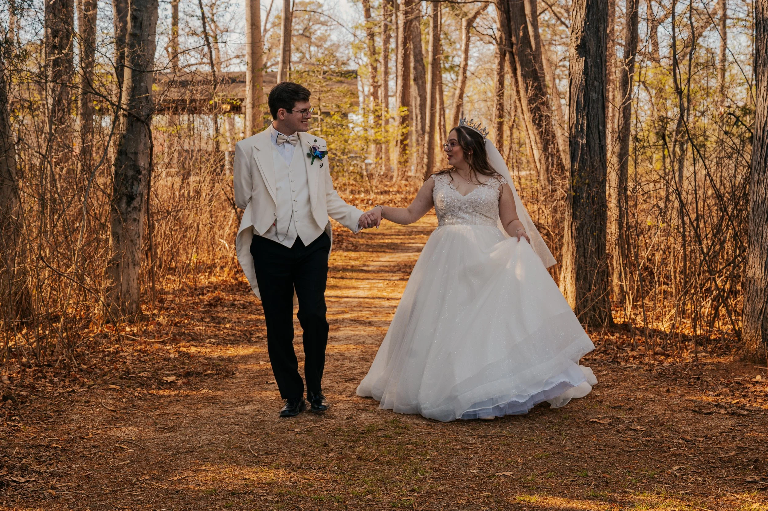 A bride and groom walking hand in hand through a wooded area during autumn, smiling at each other. The groom is in a white tuxedo with a bowtie, and the bride is in a white wedding gown with a tiara and veil.  Pennsylvania, Pittsburgh, New jersey