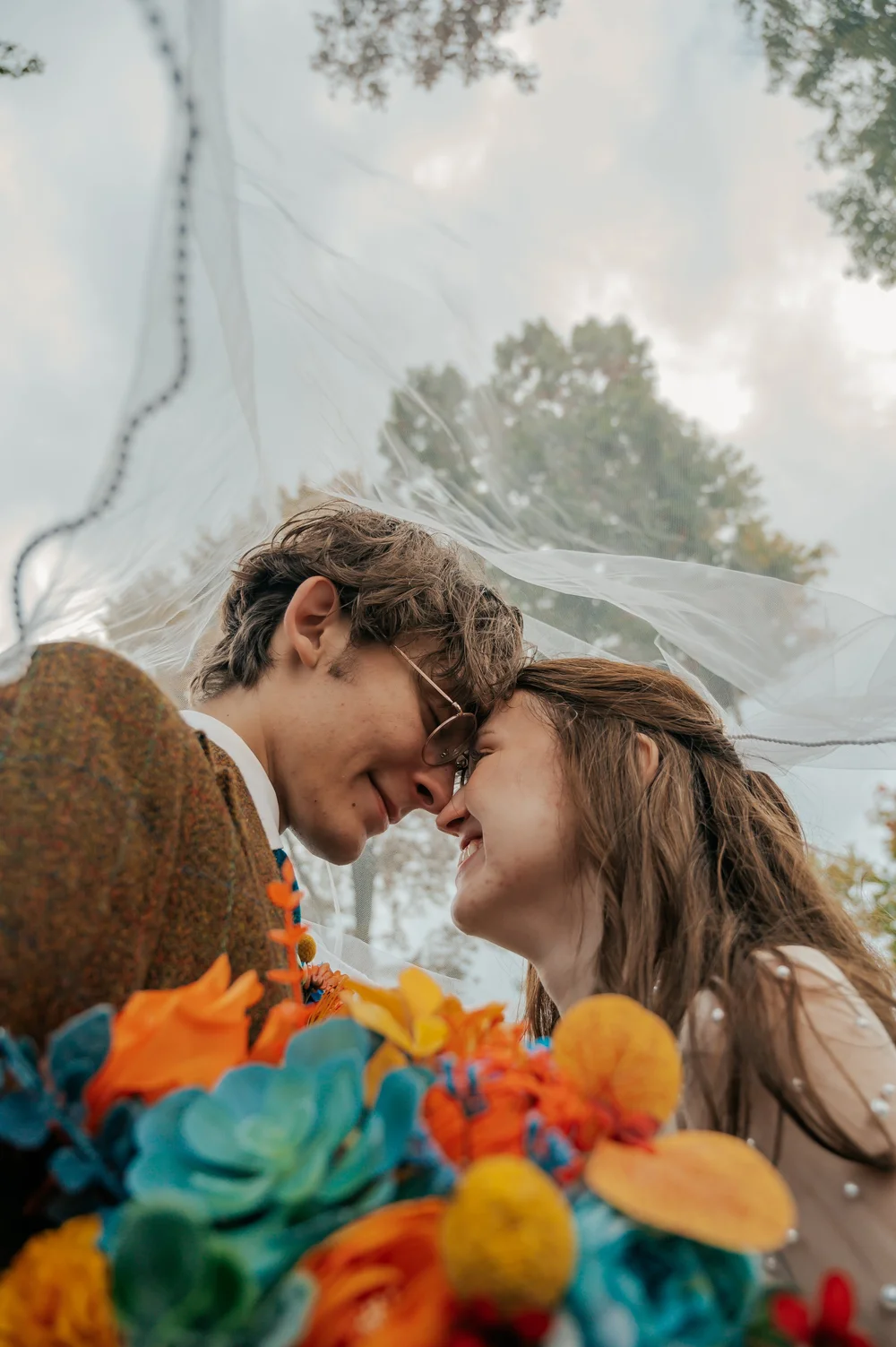 A couple smiling at each other under a veil, with colorful flowers in the foreground and cloudy sky in the background
