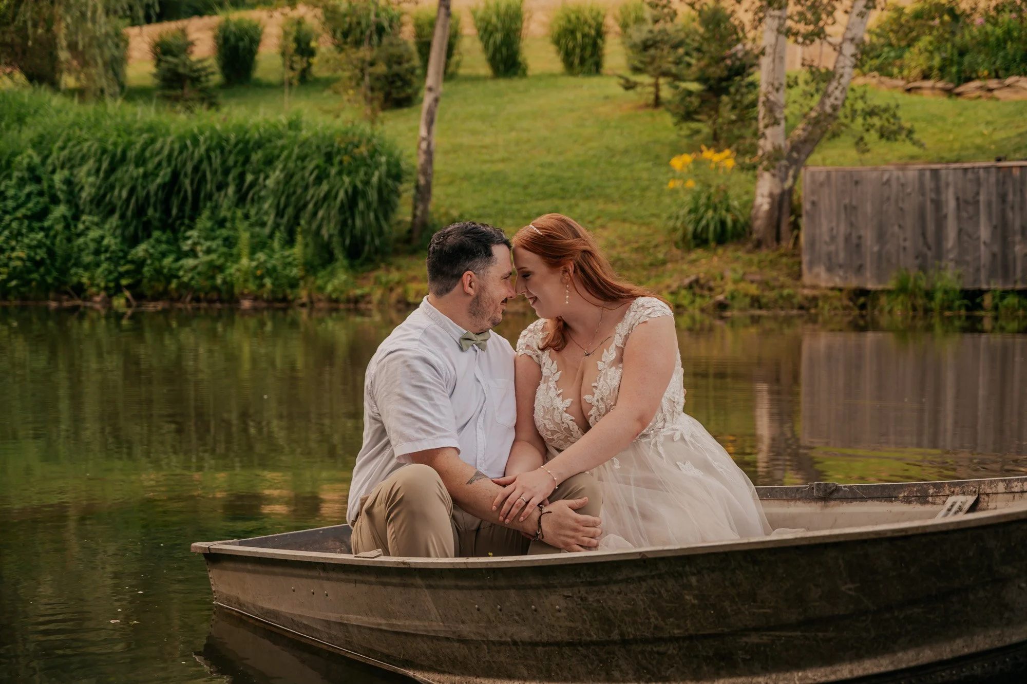 A couple in wedding attire sitting in a boat on a lake, with trees and greenery in the background.