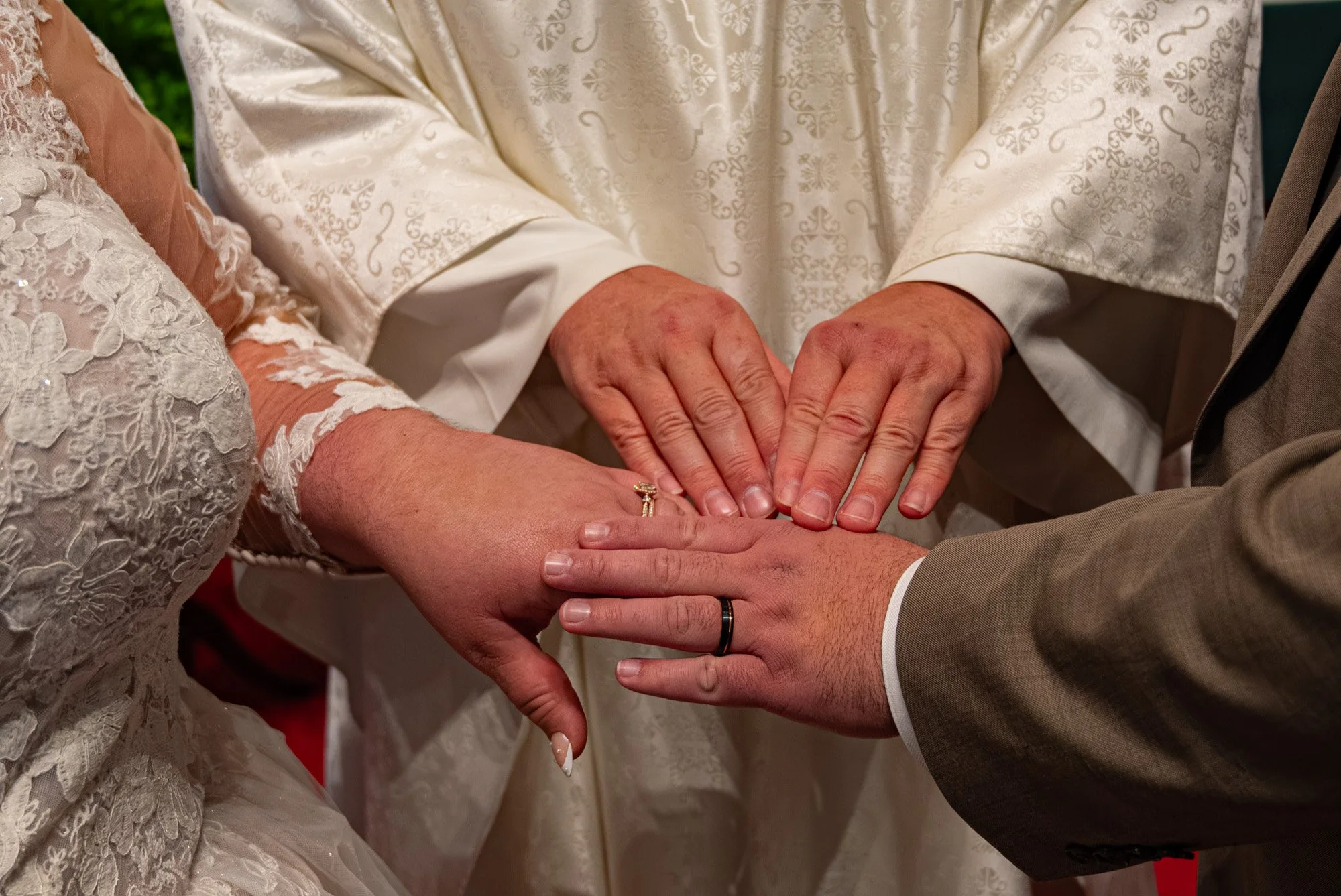 People holding hands during a wedding ceremony, with one hand wearing a wedding band. The bride in a lace dress and the groom in a beige suit are joined.