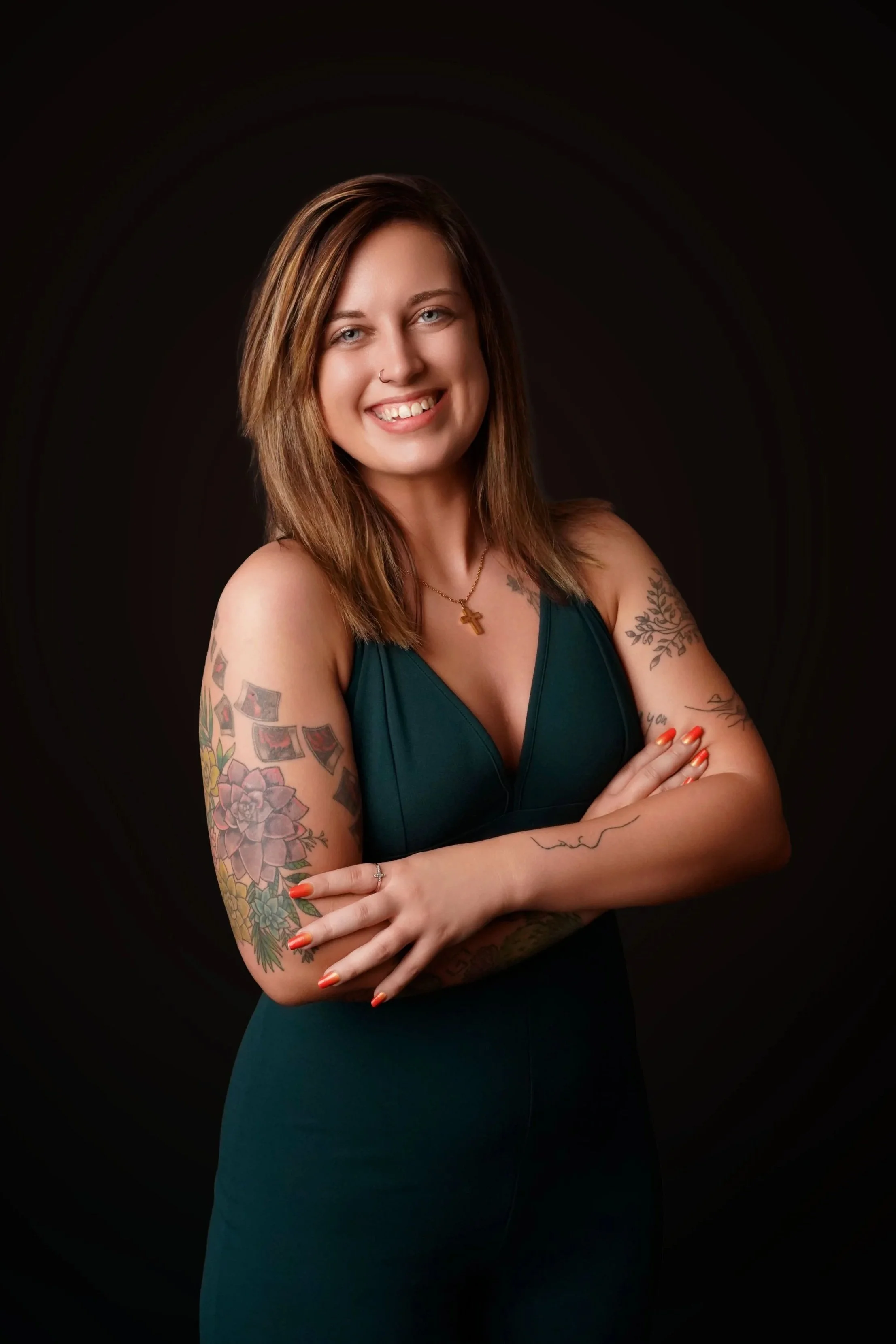 A woman with shoulder-length brown hair, wearing a dark green sleeveless top, smiling with arms crossed, standing against a black background.