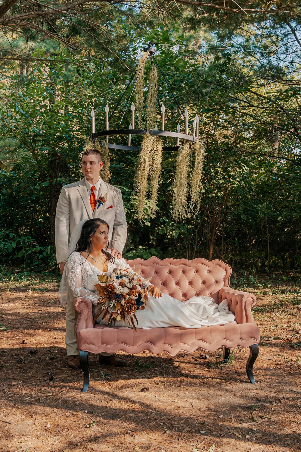 Bride on a pink sofa with bouquet, groom in a beige suit standing behind, outdoor setting with chandelier