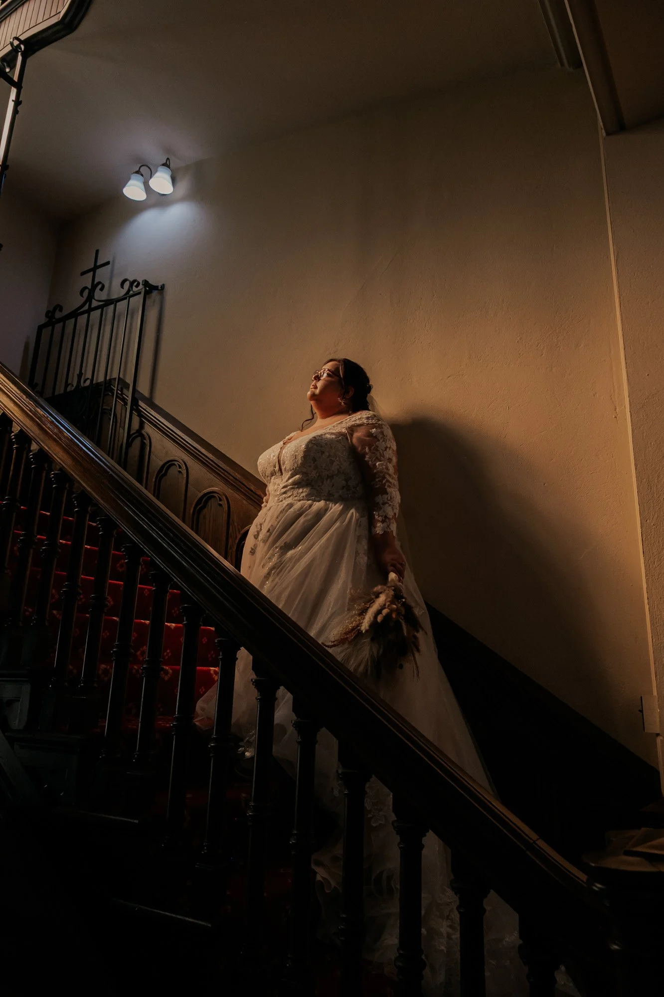 A woman in a wedding dress stands on a staircase with a wooden railing, holding a bouquet, illuminated by a ceiling light. 