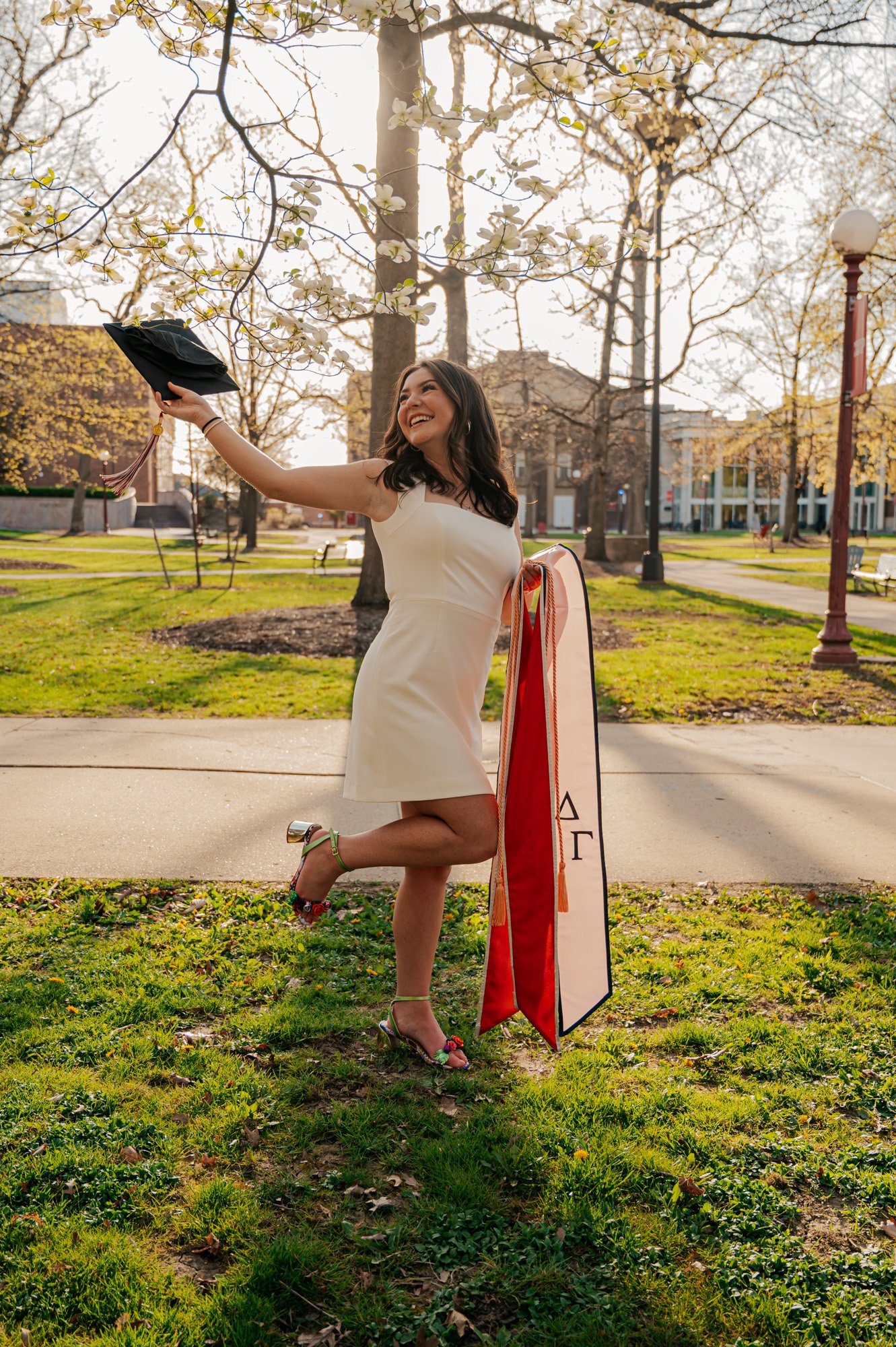 Woman in white dress holding graduation cap and sash with Greek letters, smiling outdoors on a sunny day.