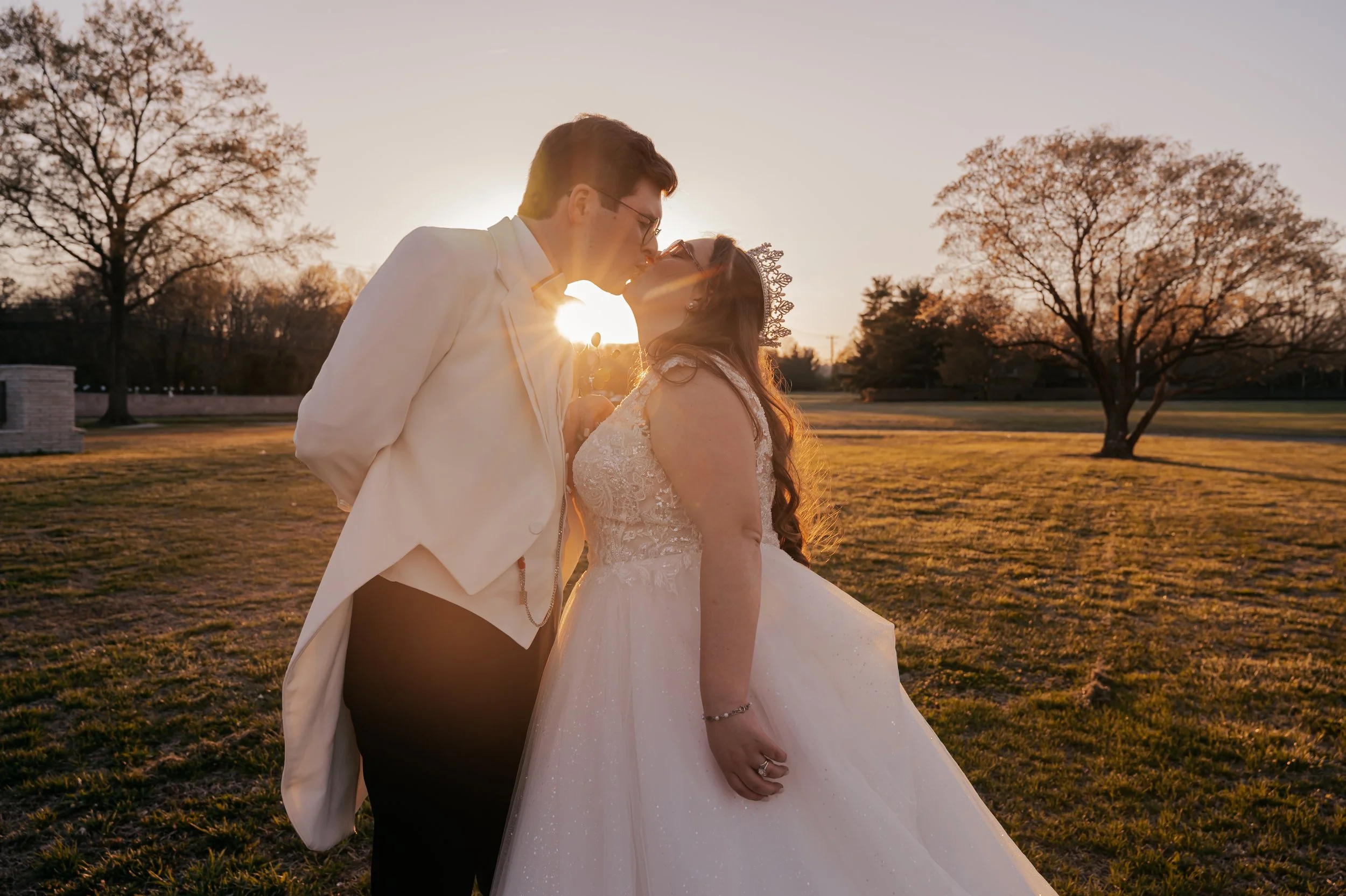A newlywed couple sharing a kiss outdoors during sunset, with trees in the background and the sun creating a warm glow around them.  Johnstown Pennsylvania, Pittsburgh, New Jersey, elopement Photographer, elope