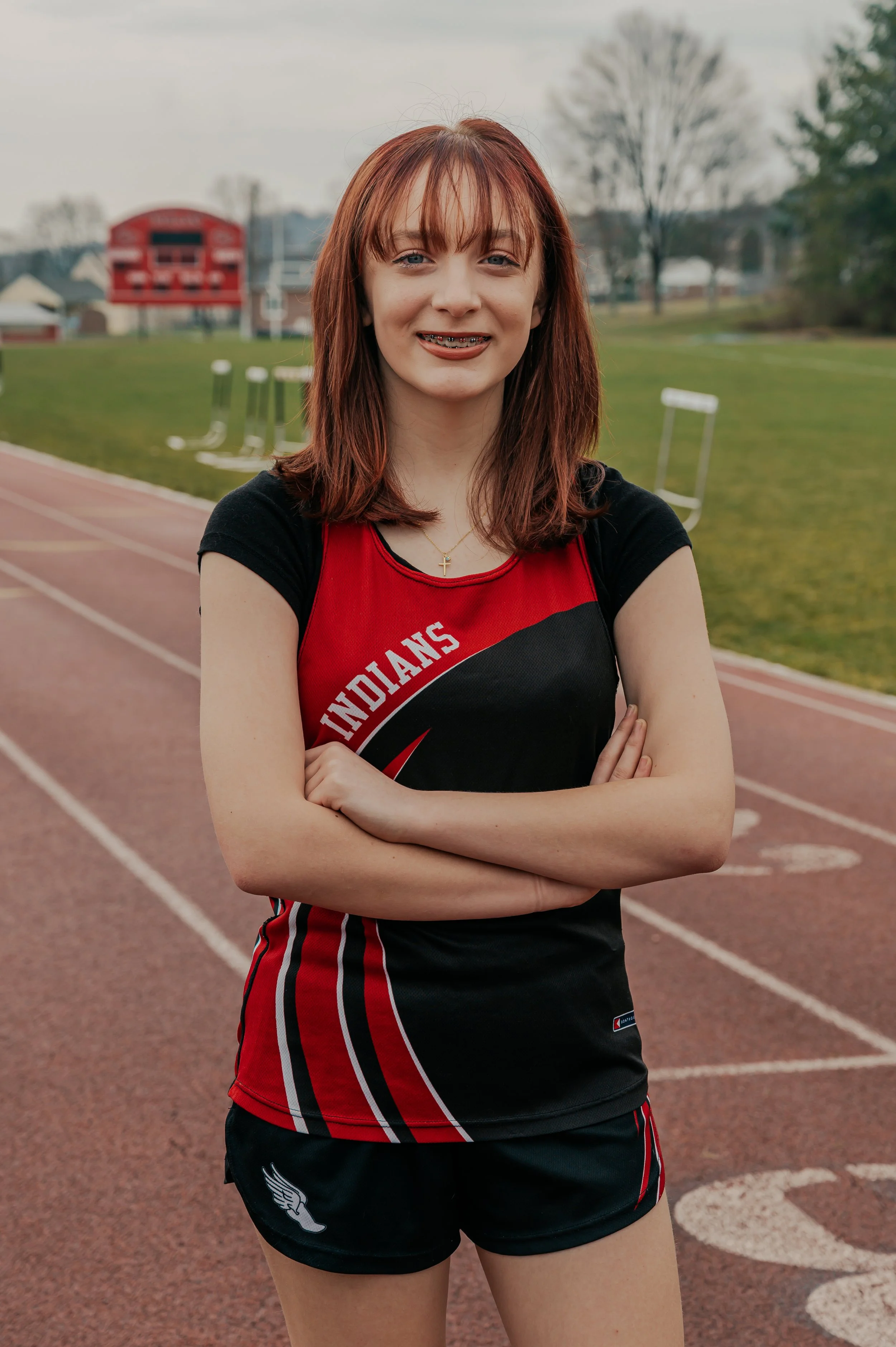 A young woman with shoulder-length red hair and braces, standing on a running track with her arms crossed, wearing a black and red track uniform with the word 'Indians' on the front.