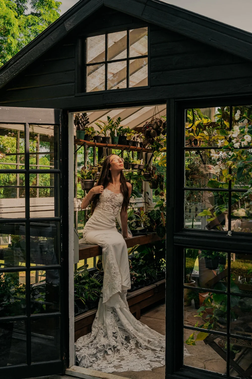 A woman in a white wedding dress sitting inside a greenhouse filled with plants, with sunlight coming through the glass roof