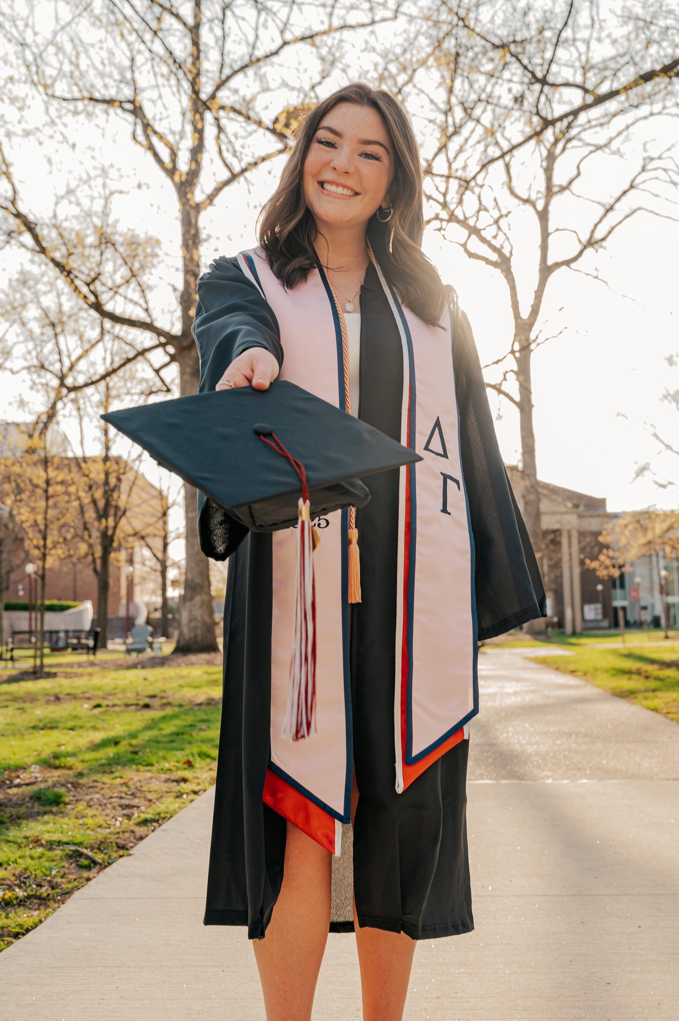 A female graduate in a cap and gown holding out her graduation cap outdoors on a sunny day.