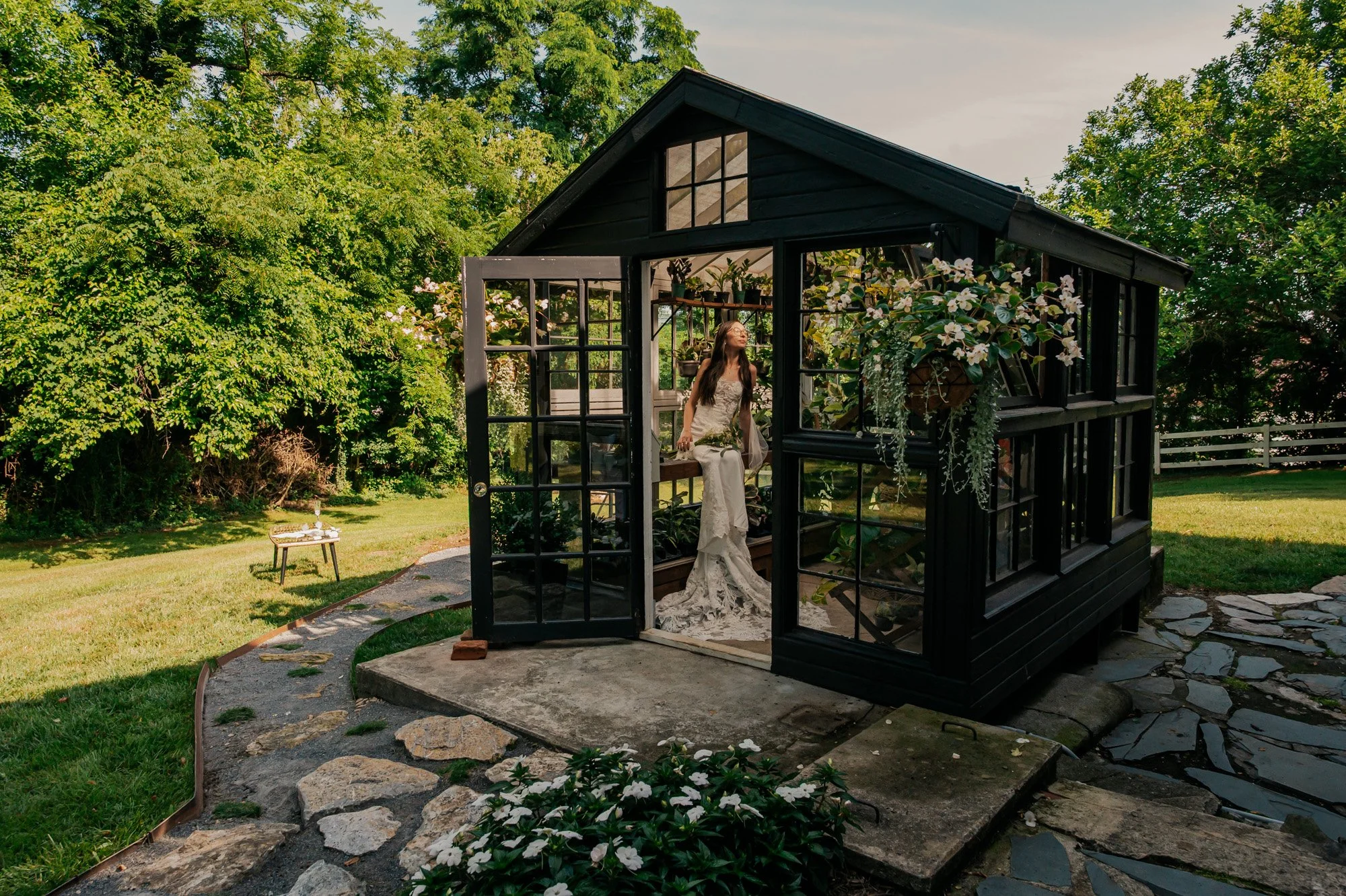 A woman in a wedding dress sitting inside a black garden greenhouse with open glass doors, surrounded by greenery and flowers, during early evening.
