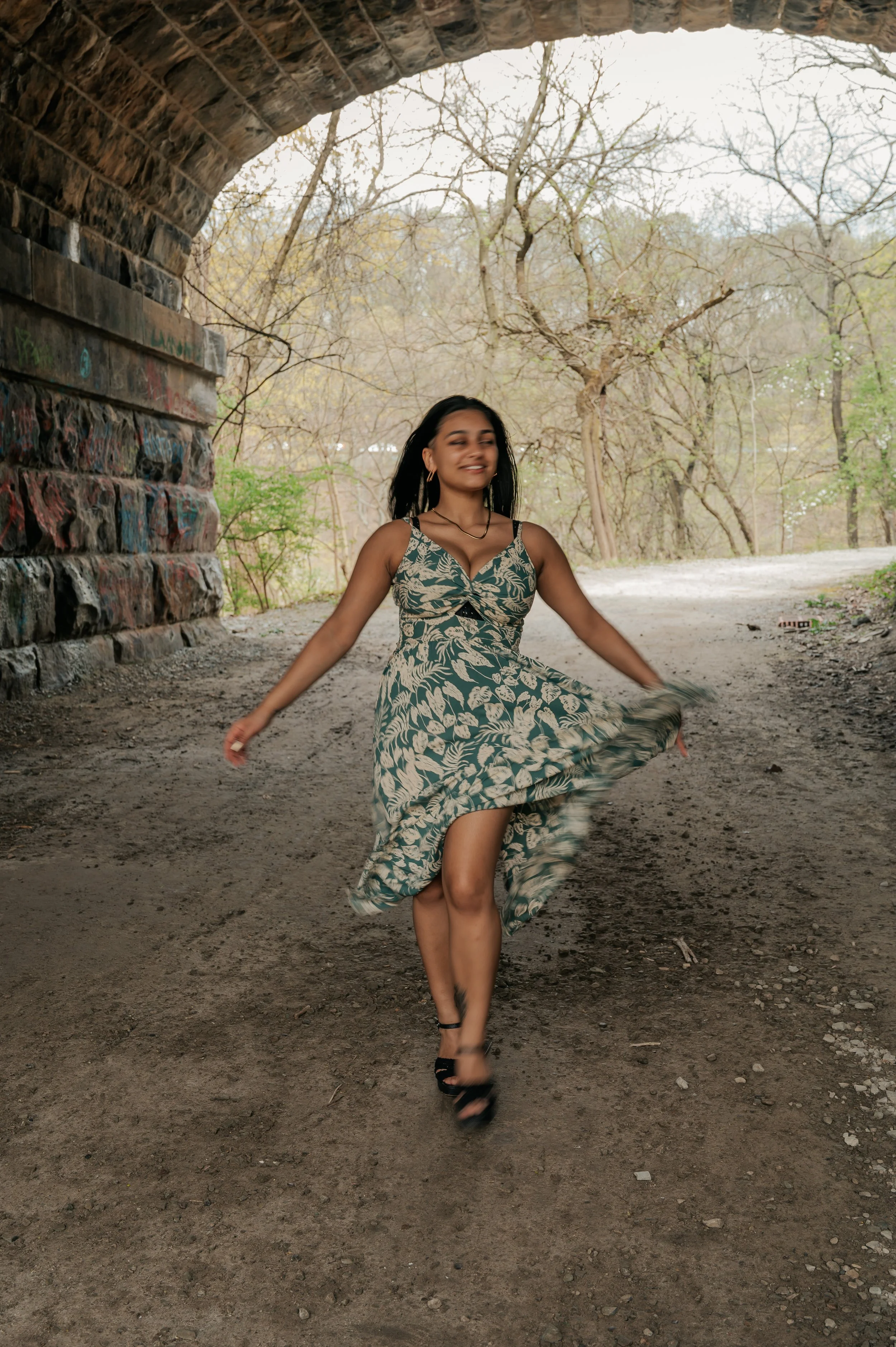 A woman in a floral dress twirling under a bridge on a dirt path in a park with leafless trees in the background.