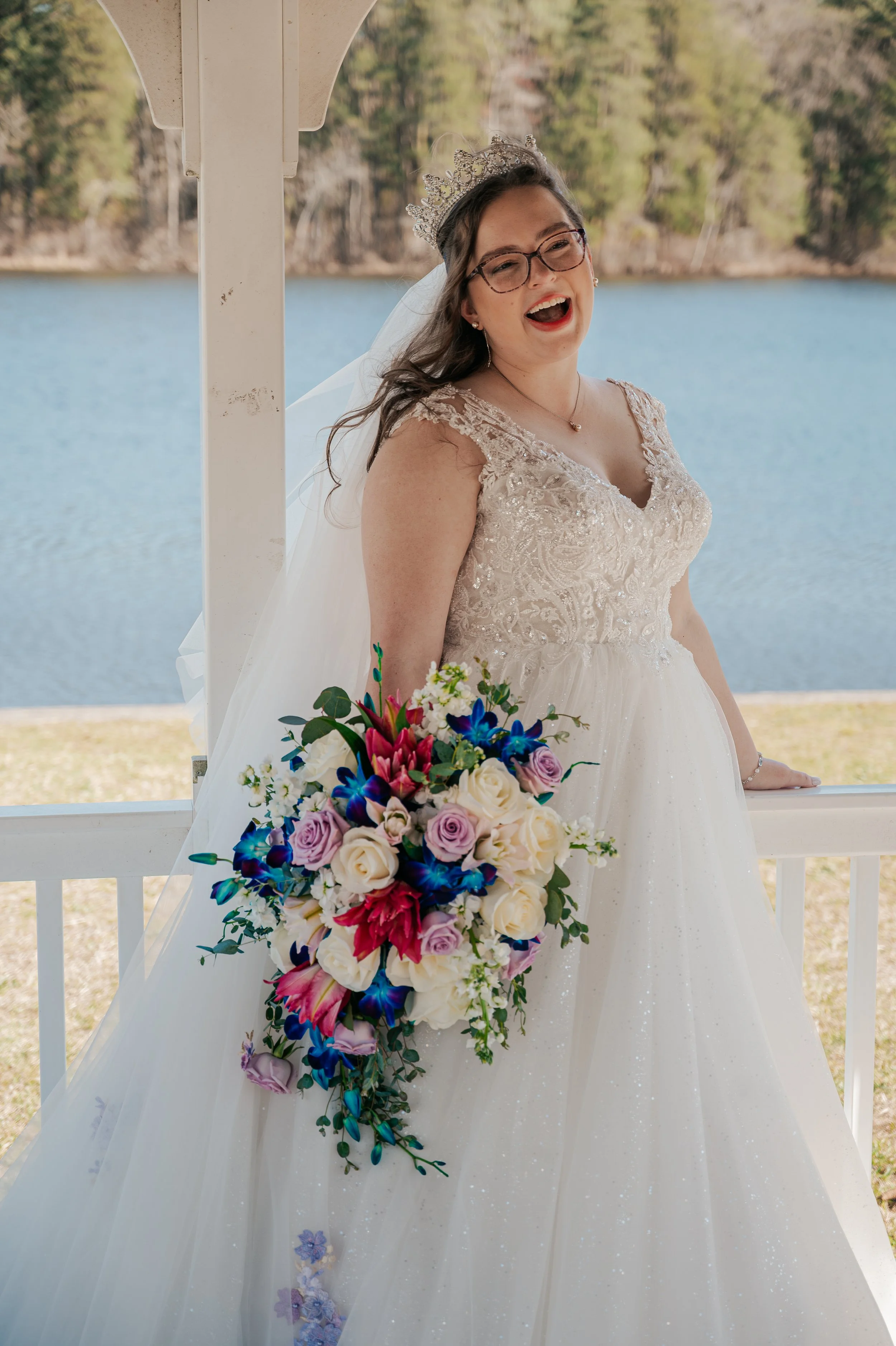A smiling woman in a wedding dress with glasses and a tiara, holding a colorful bouquet of flowers, standing outdoors near a lake and trees.
Johnstown Pennsylvania, Pittsburgh, New Jersey, elopement Photographer, elope