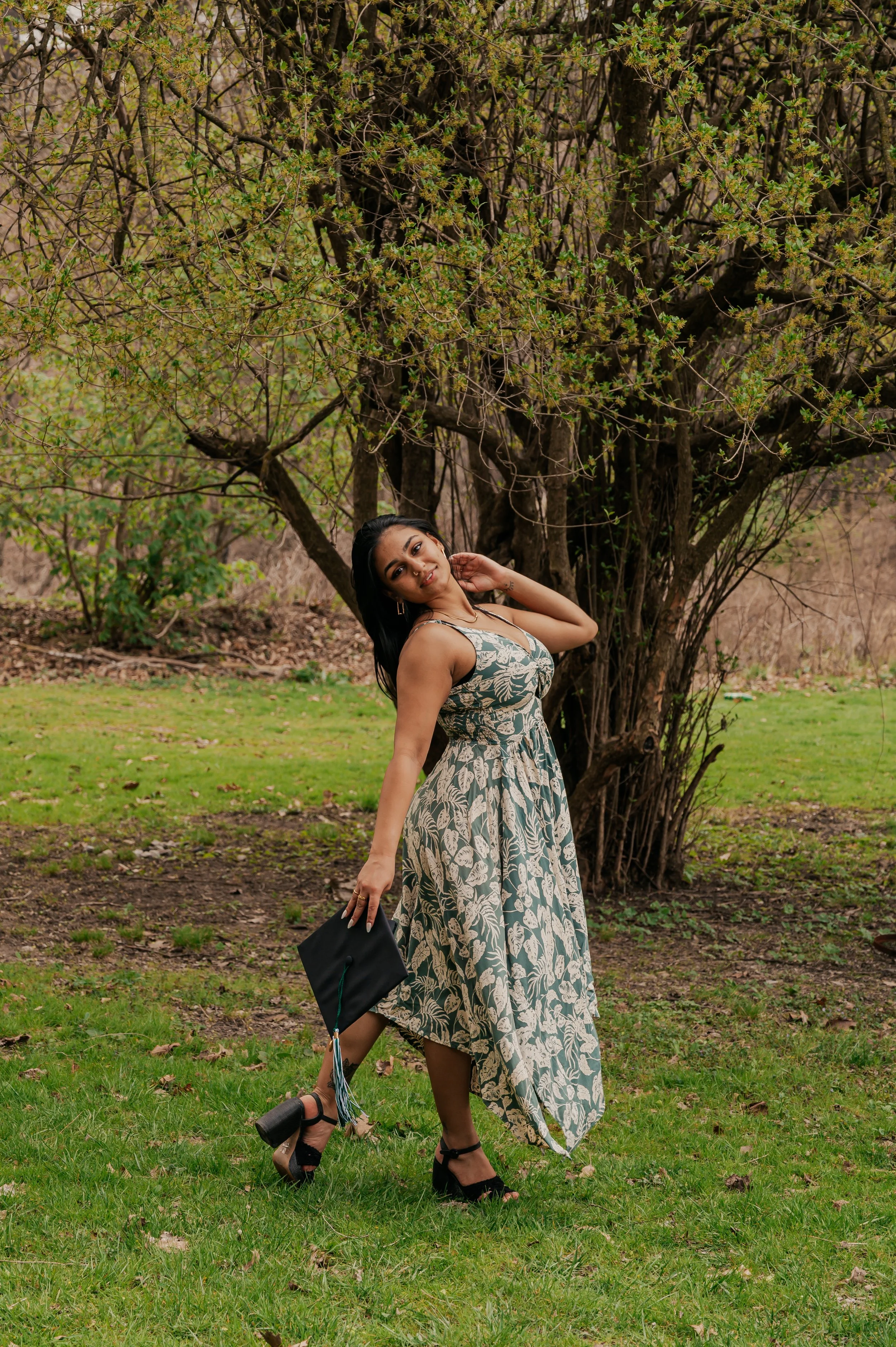 A woman in a patterned dress holding a graduation cap and posing outdoors near a tree with green leaves.