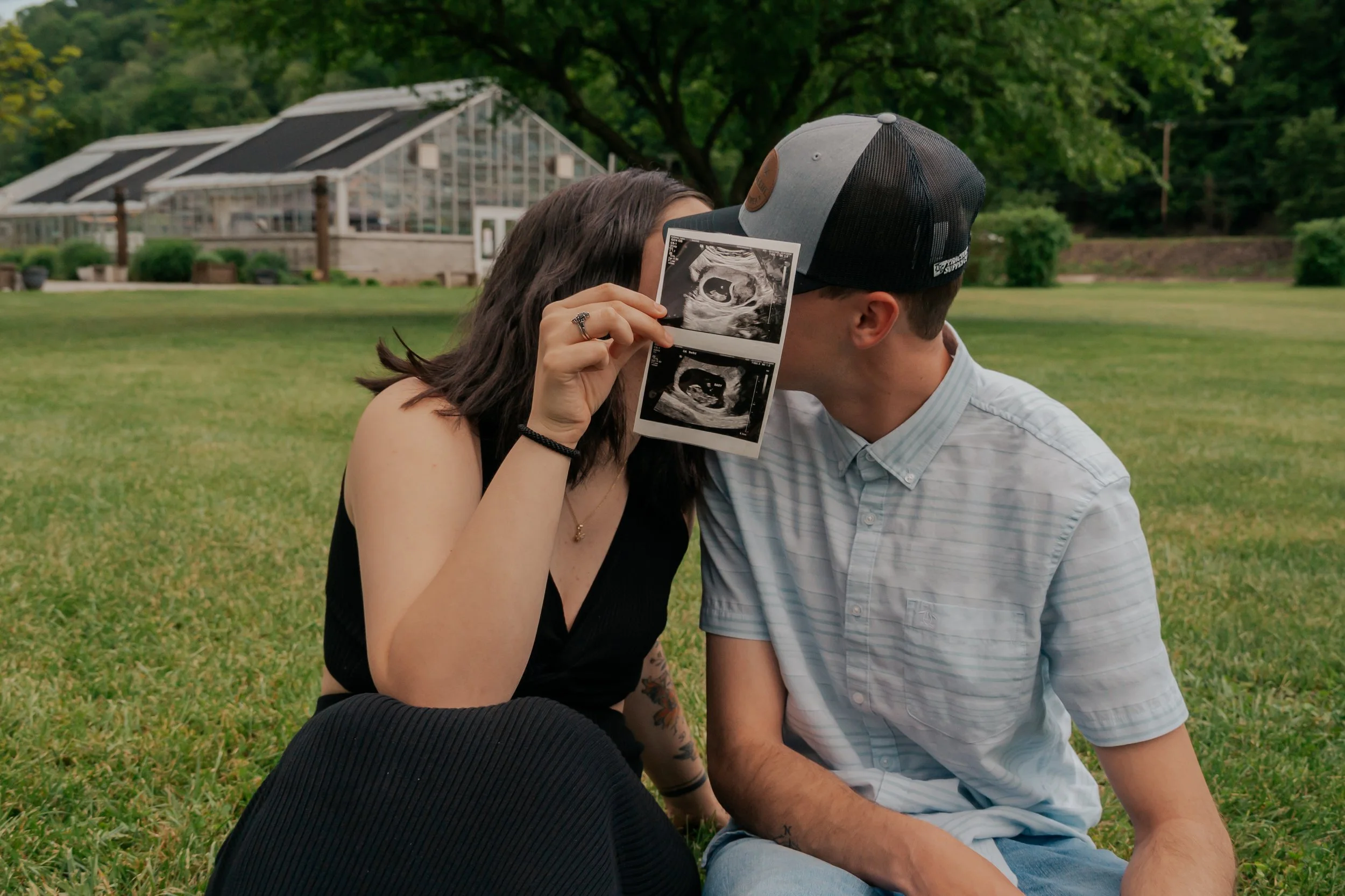 A couple sitting on grass outdoors, sharing an ultrasound image, with a greenhouse and trees in the background.