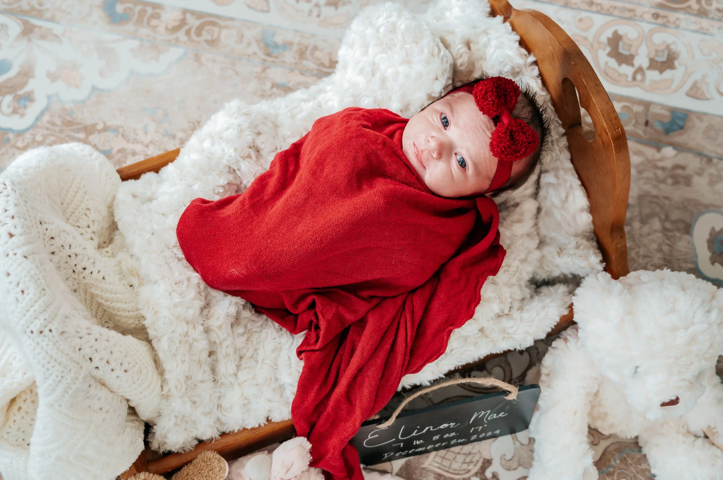 A newborn baby girl wrapped in a red blanket, lying on a fluffy white blanket on a small wooden bed, wearing a red headband with a bow, with a knitted cream-colored blanket and a stuffed animal nearby.