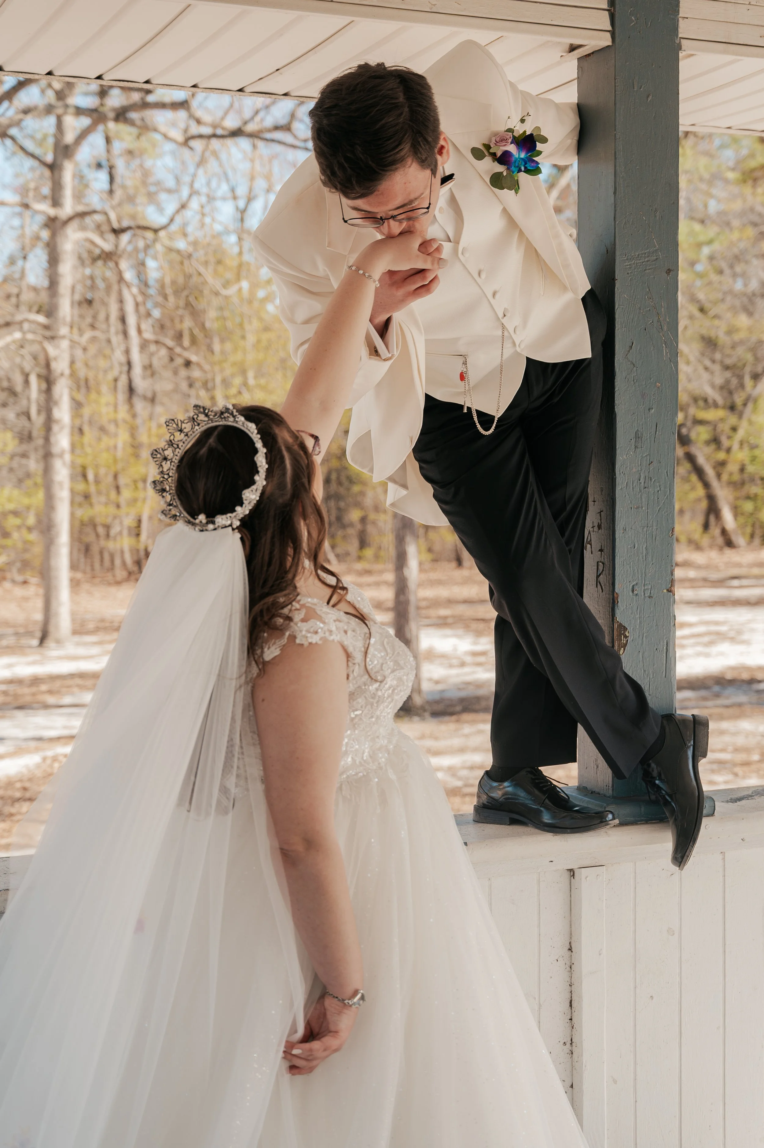 A groom in a white tuxedo kissing a bride who is wearing a wedding dress and a tiara in a rustic outdoor setting with trees in the background. Johnstown Pennsylvania, Pittsburgh, New Jersey, elopement Photographer, elope