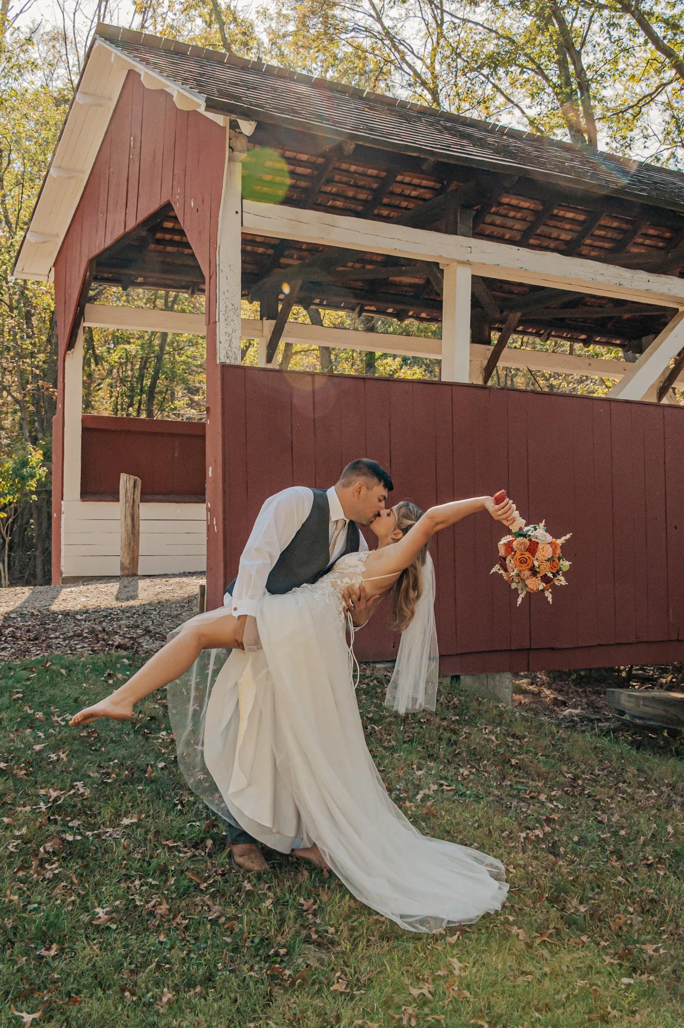 A bride and groom share a kiss outdoors, with the groom holding the bride in a dip. The bride is holding a bouquet of roses and peonies. The background features a small red and white wooden building, surrounded by trees with autumn leaves.