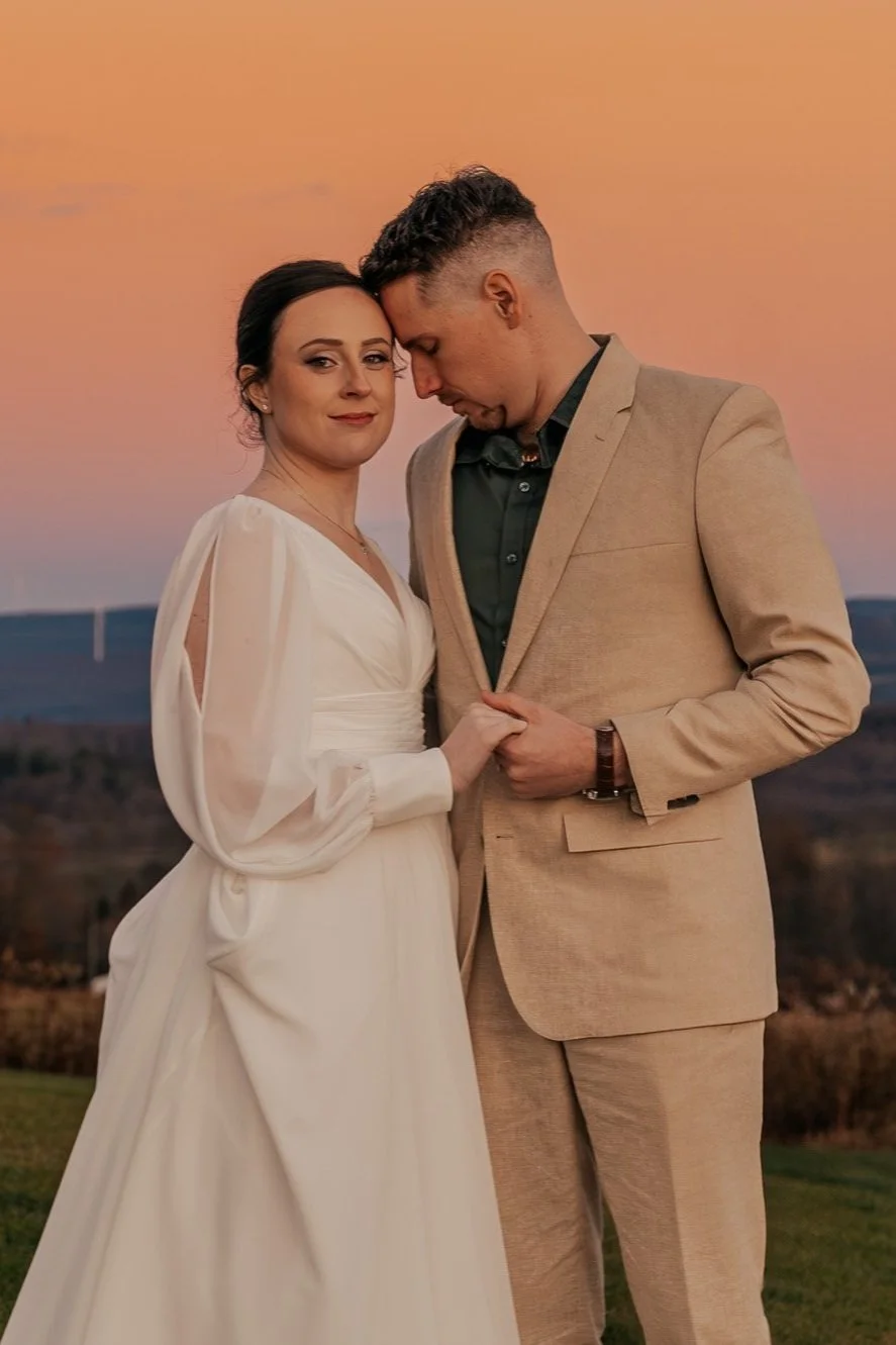 A couple embraces at sunset, with the woman in a white dress and the man in a beige suit
