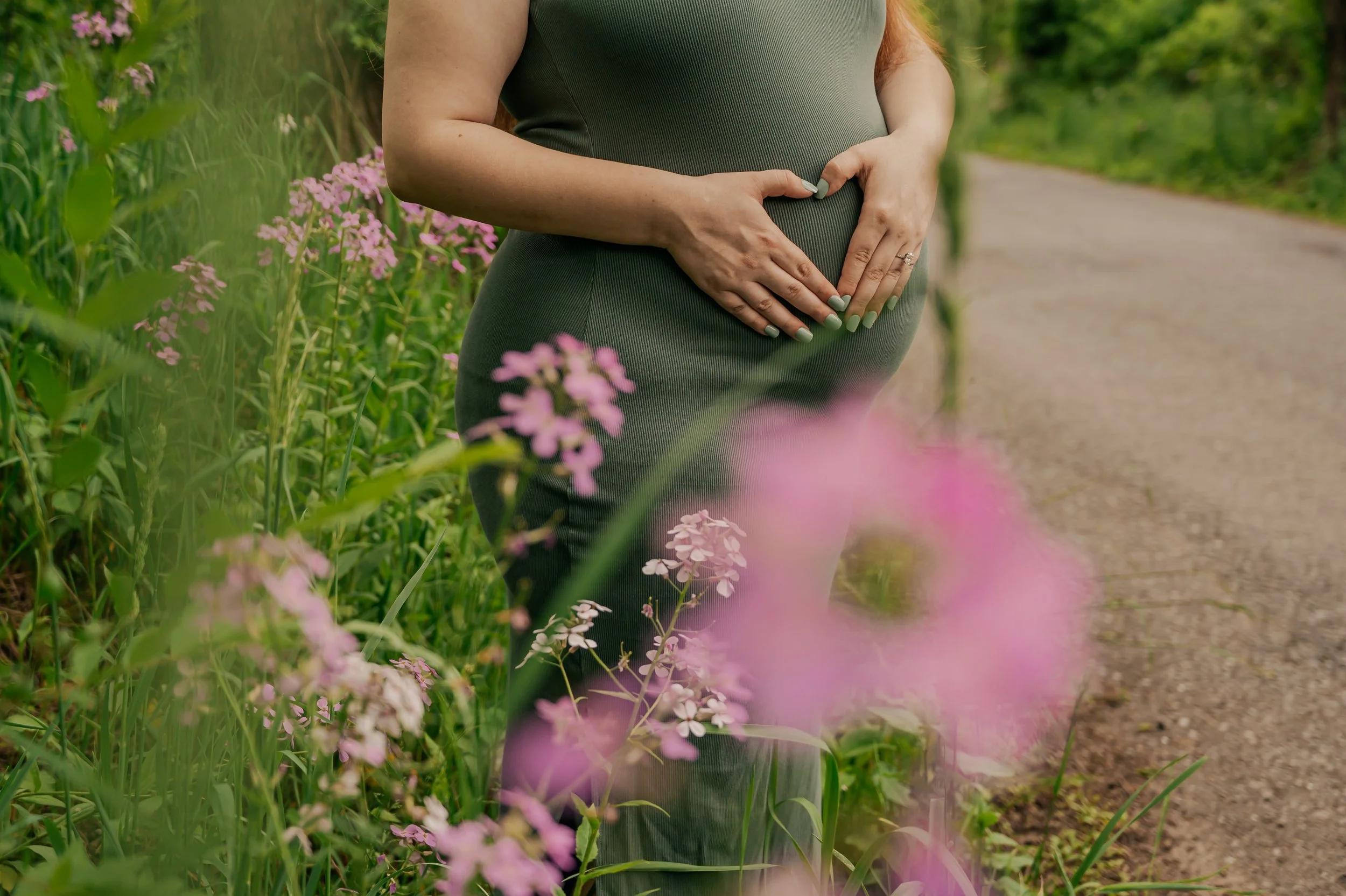 Pregnant woman standing by a dirt path in a green field with pink flowers, holding her belly with both hands.