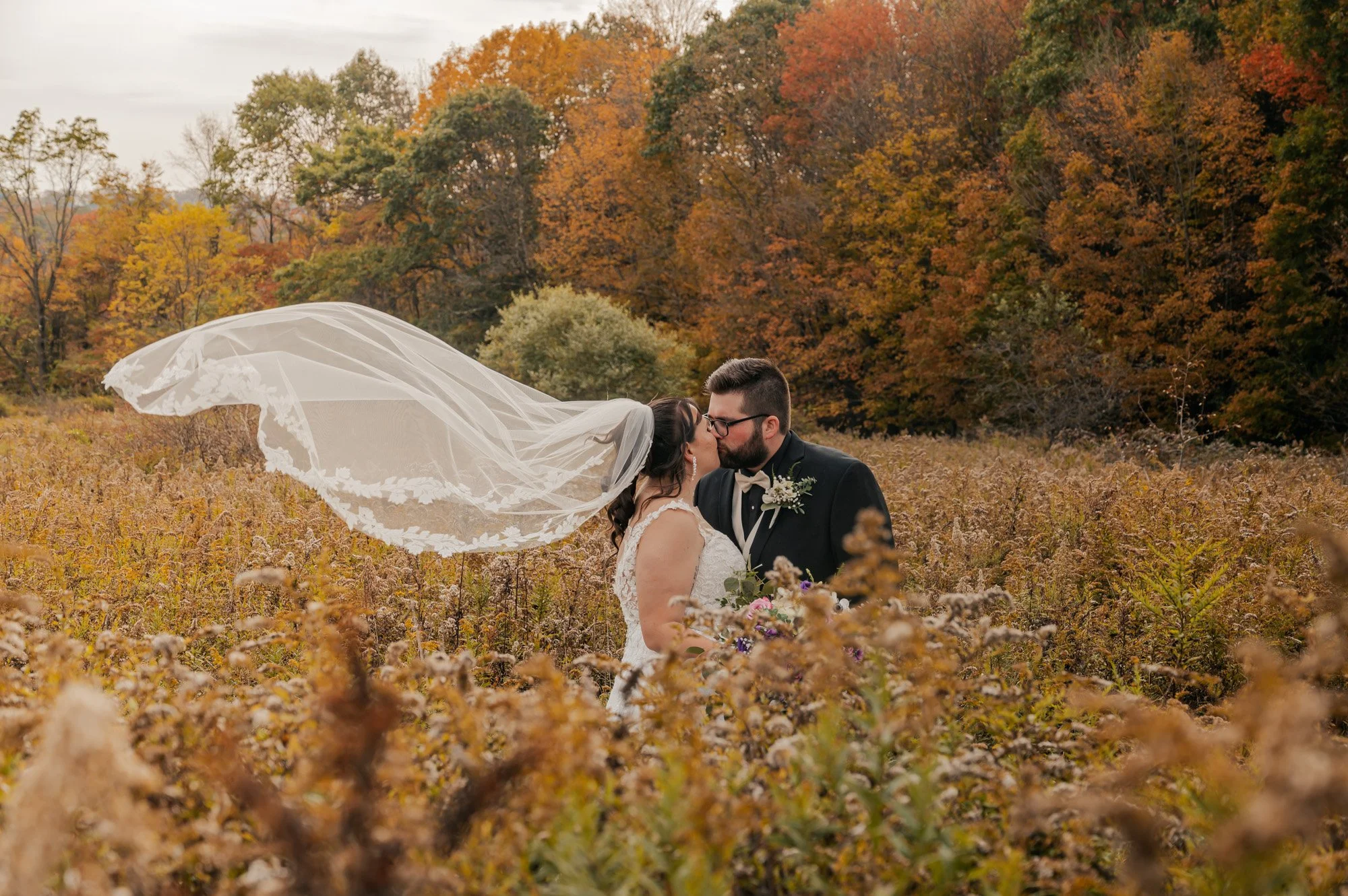 Bride and groom kissing in autumn field with flowing veil, surrounded by fall foliage.