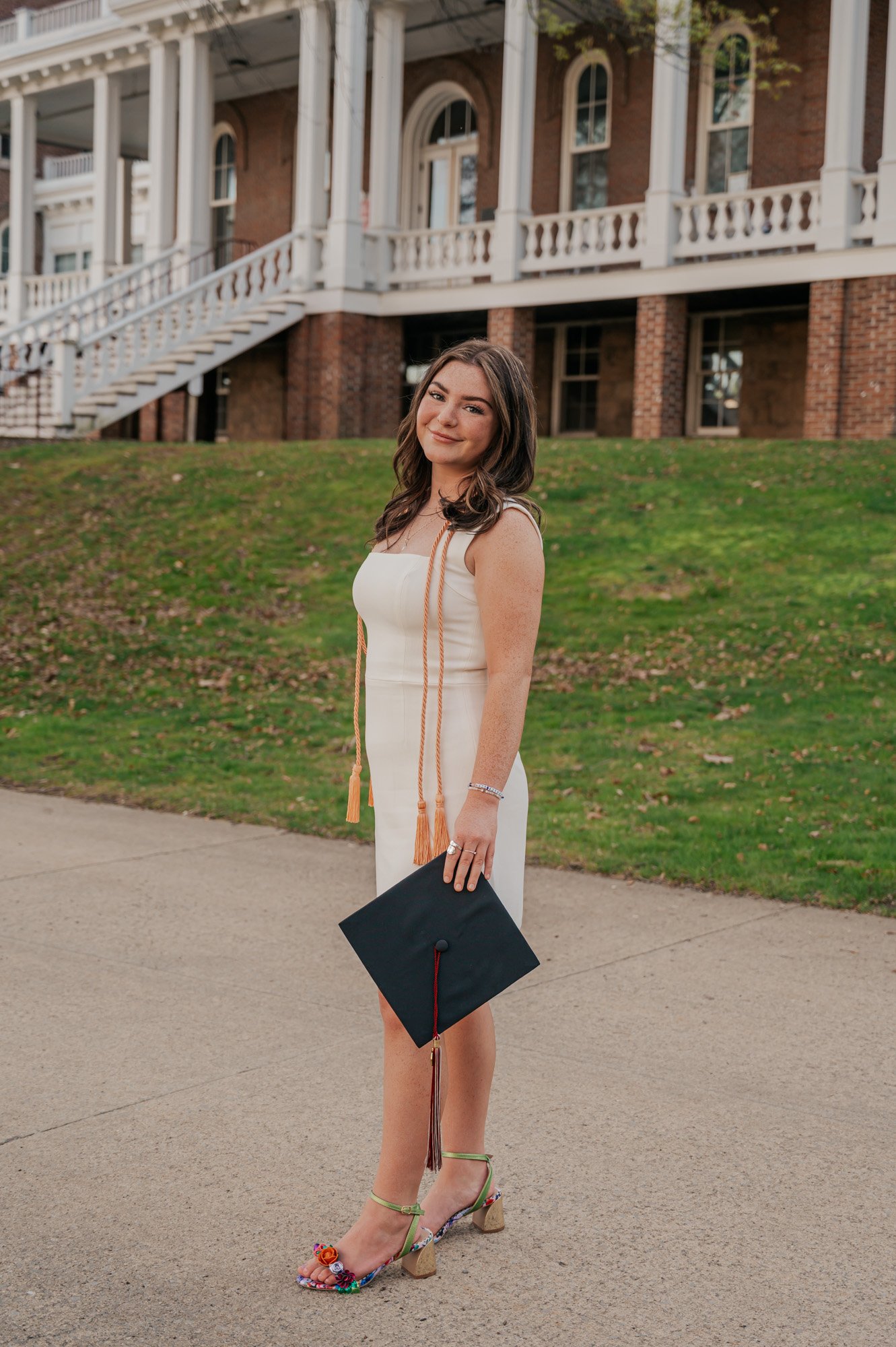Young woman in white dress holding a graduation cap, standing on a path with a historic building in the background.