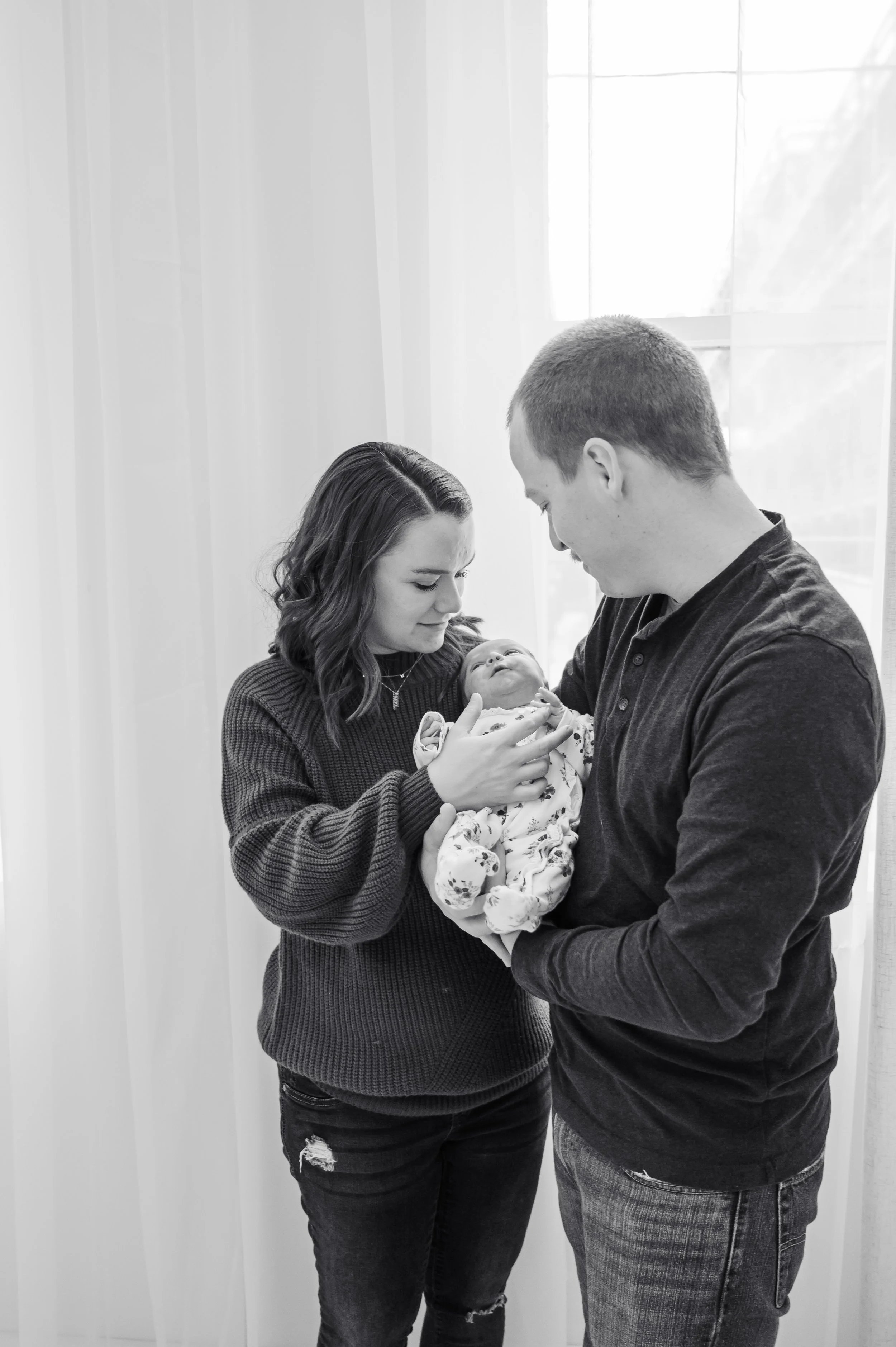 A young woman and man holding a newborn baby, looking at the baby affectionately inside a room with natural light.