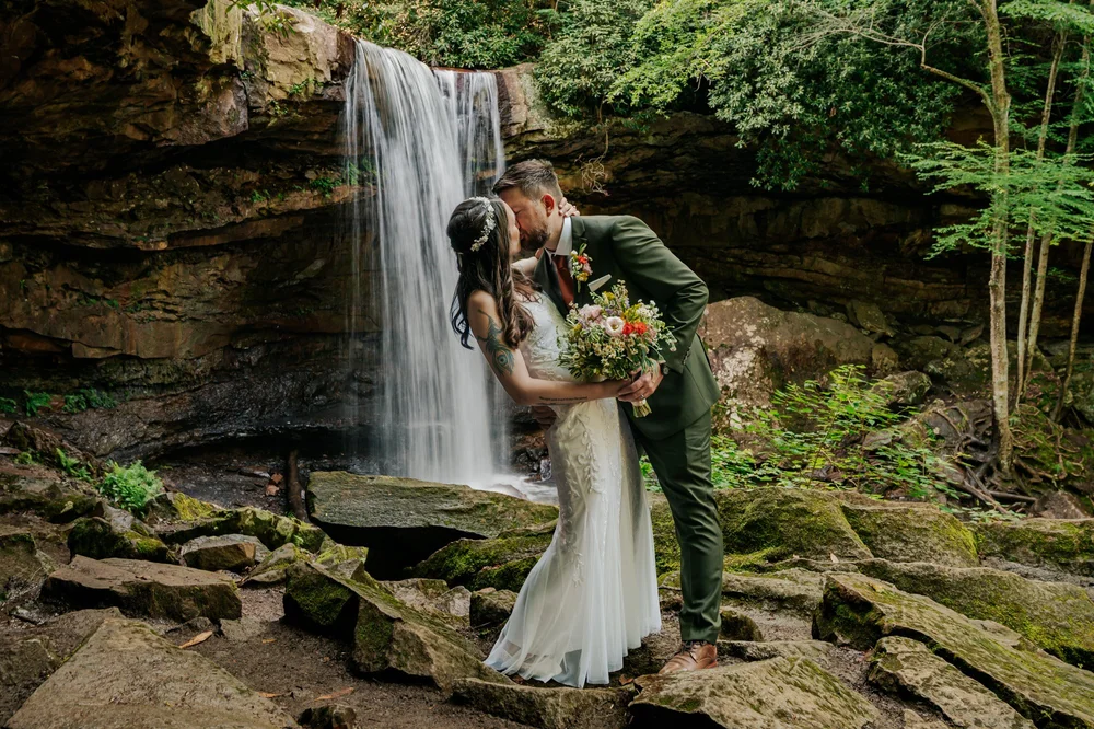 Bride and groom kissing at a waterfall, surrounded by wedding flowers