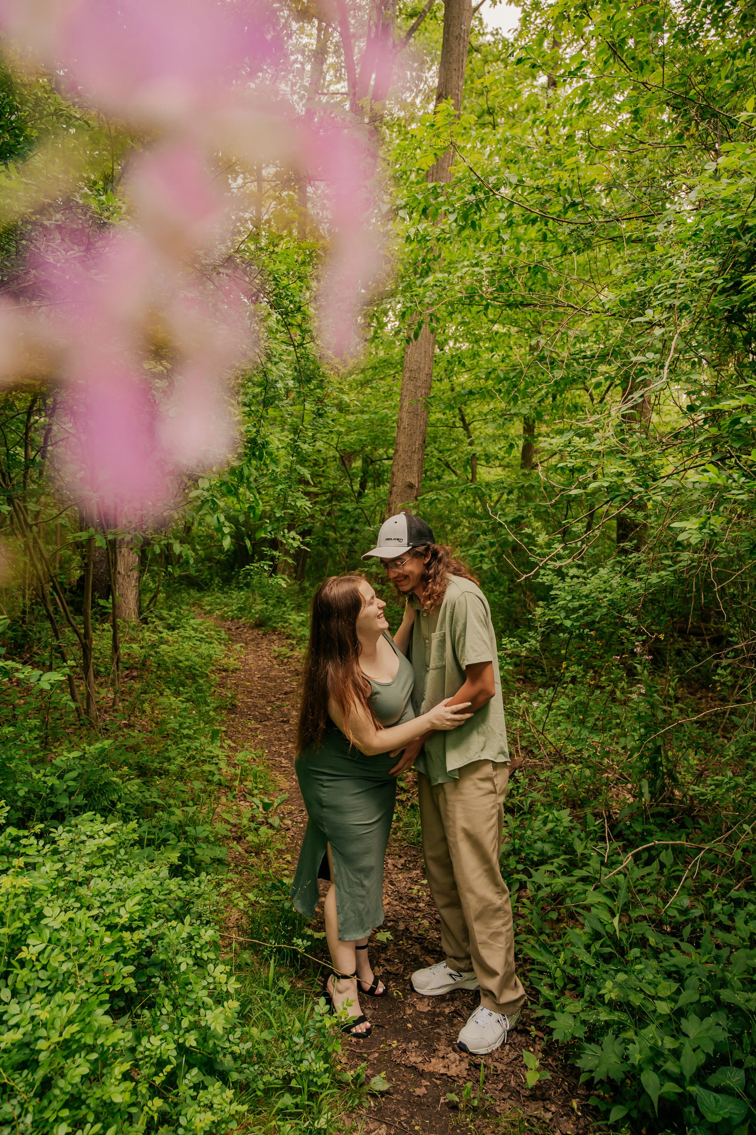 Two women standing close in a lush green forest, smiling and holding each other on a dirt path, with pink flowers blurred in the foreground.